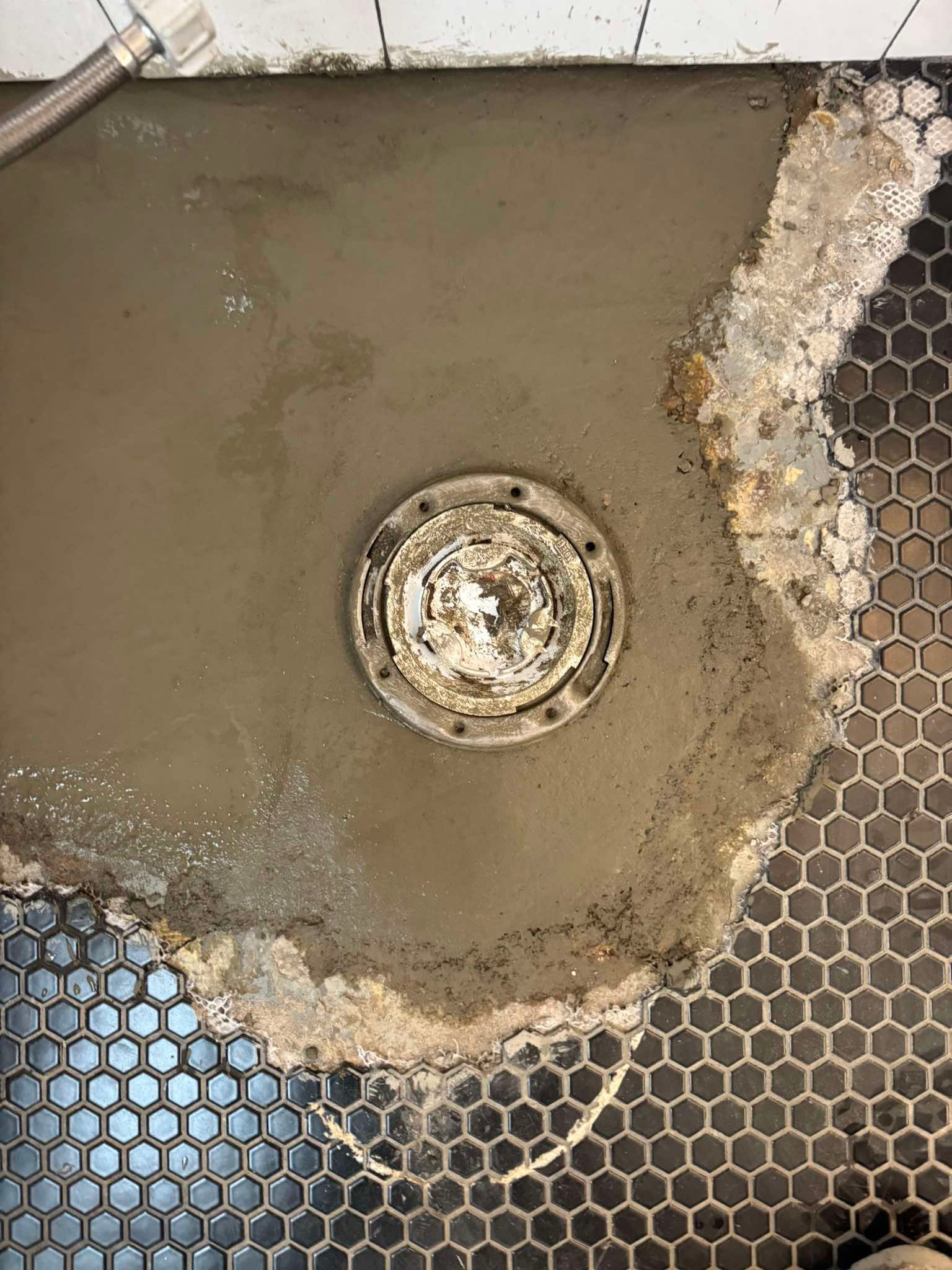 A shower drain flange surrounded by fresh cement patch work, bordered by black hexagonal floor tiles.