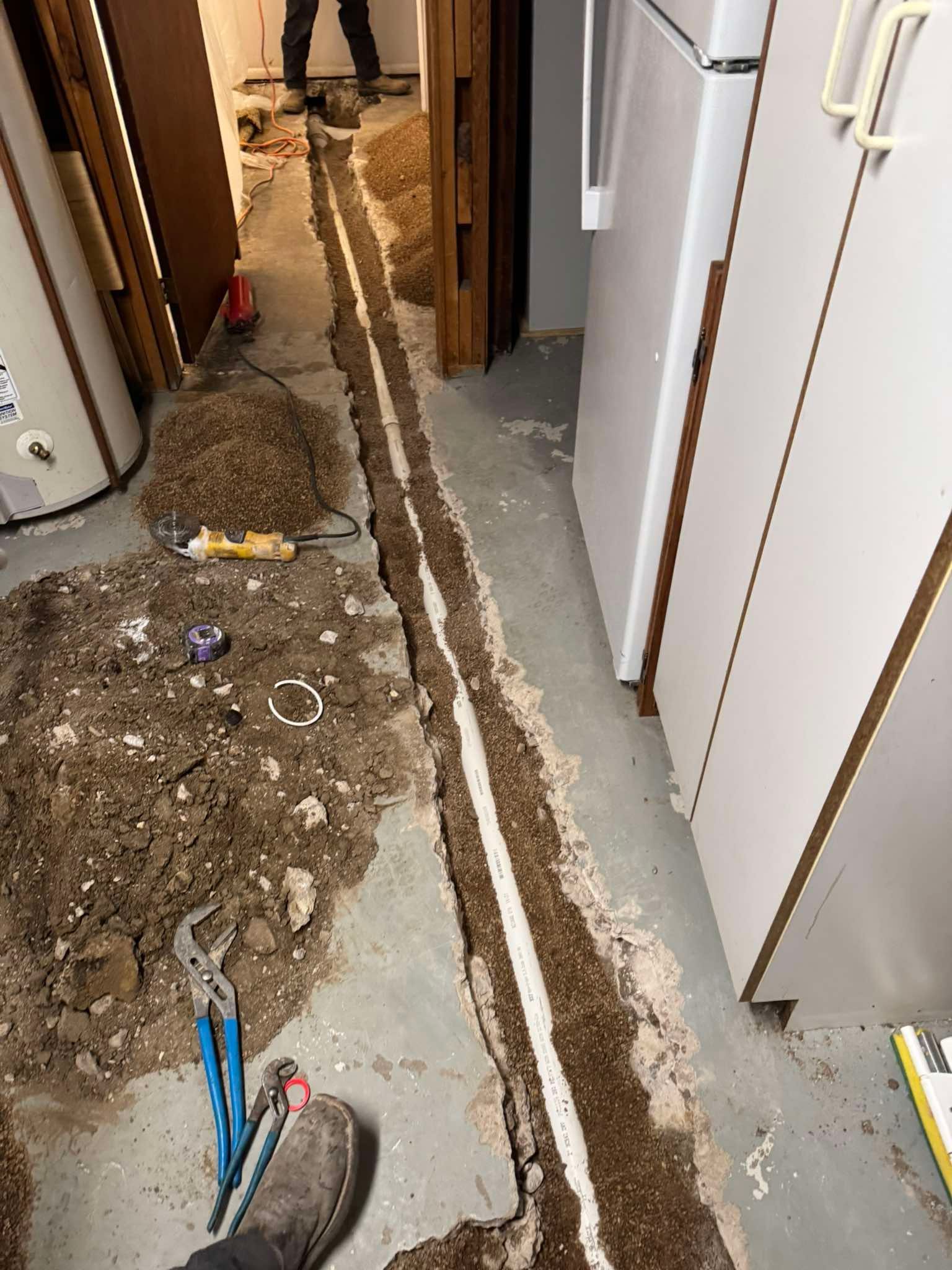 A trench cut into a concrete floor shows a newly installed white pipe in a room with a water heater and cabinets.