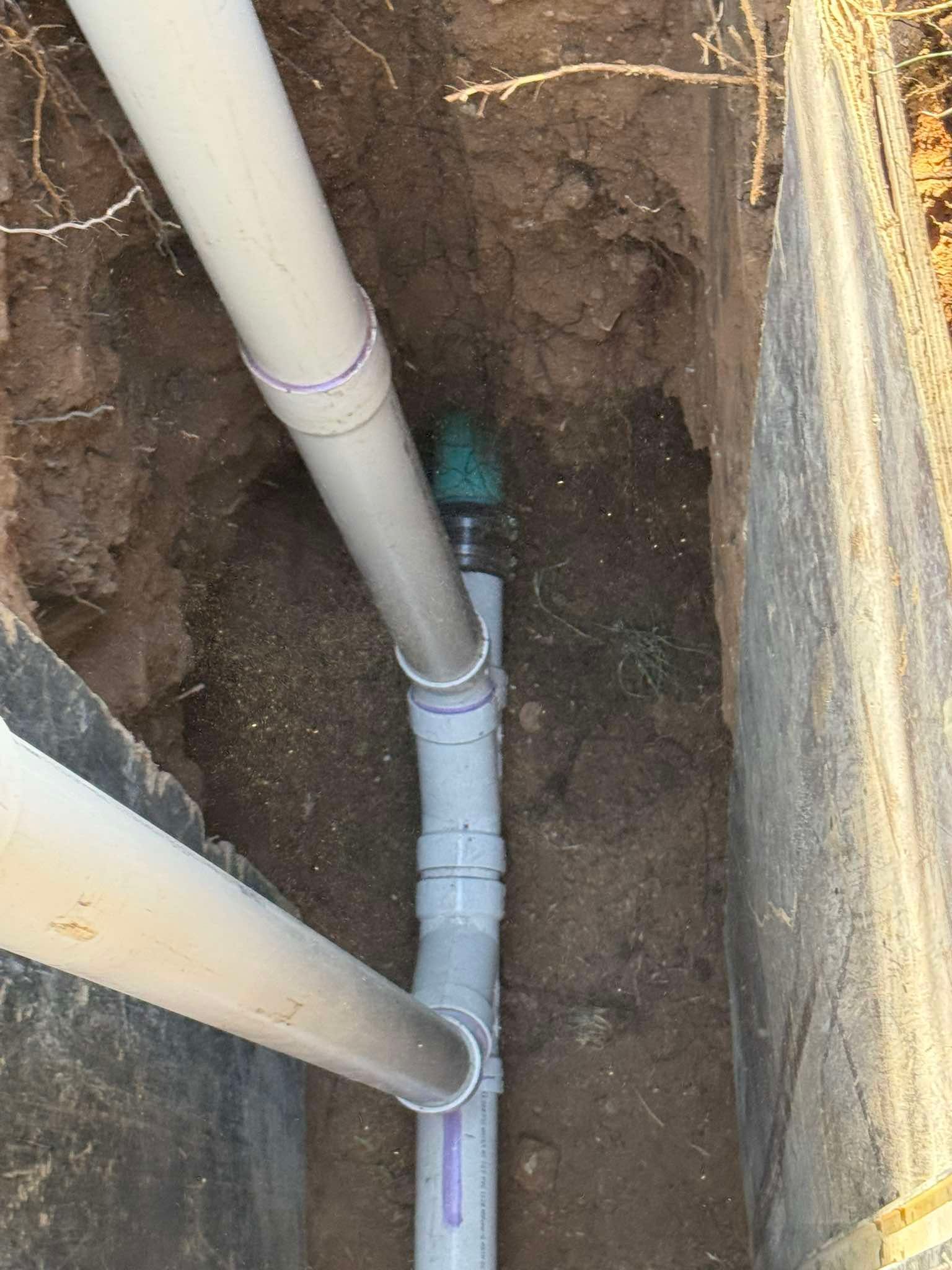 White PVC pipes connected with fittings buried in a narrow dirt trench alongside a concrete foundation wall.