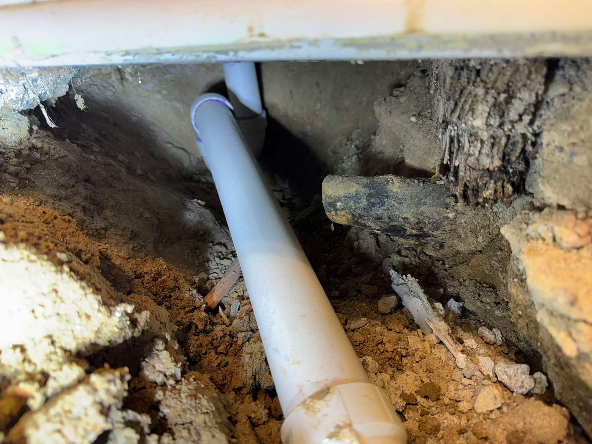 A gray PVC pipe runs through a dark, narrow gap between uneven dirt, debris, and a concrete ledge.