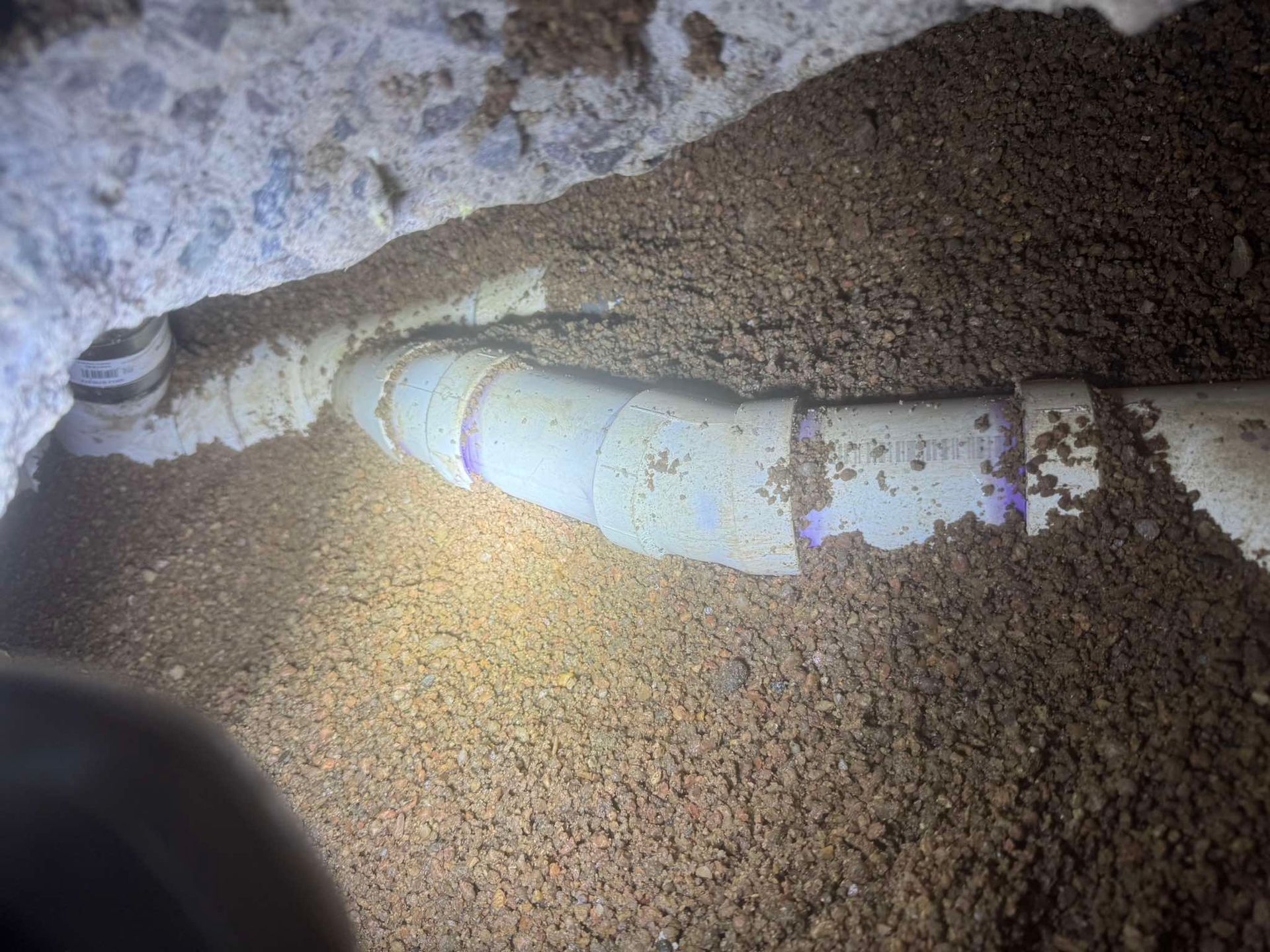 A PVC pipe with purple-stained joints partially buried in gravel under a concrete slab.