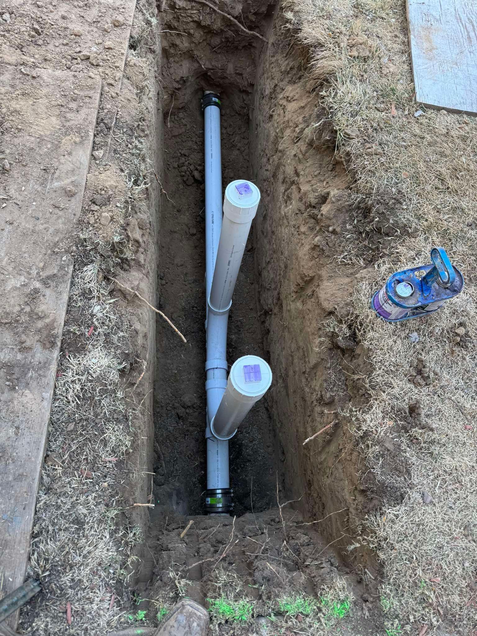 A trench in the ground containing grey PVC pipes with two vertical capped branches for plumbing access.