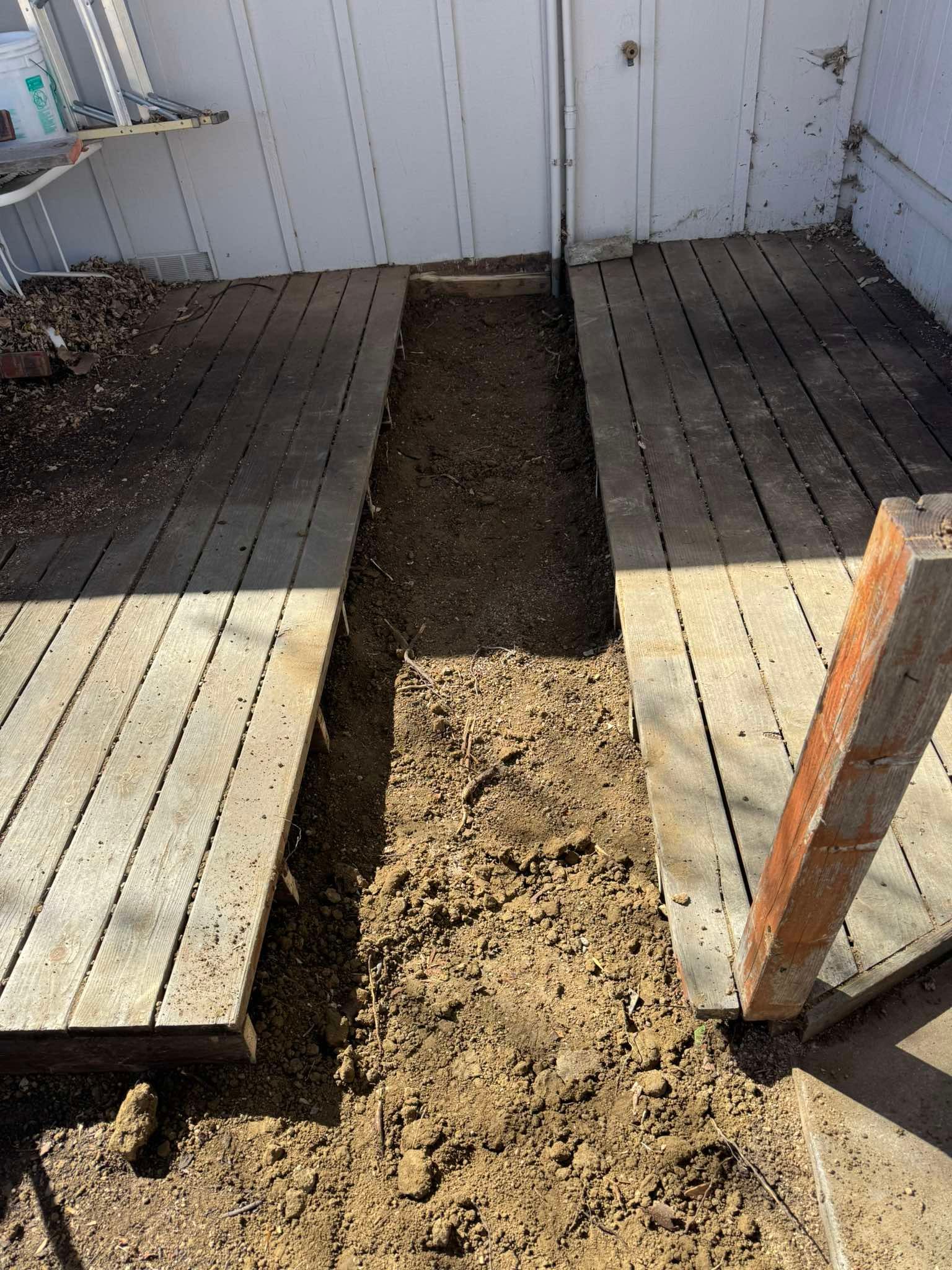 A narrow dirt trench lies between two wooden deck sections alongside a white wall.