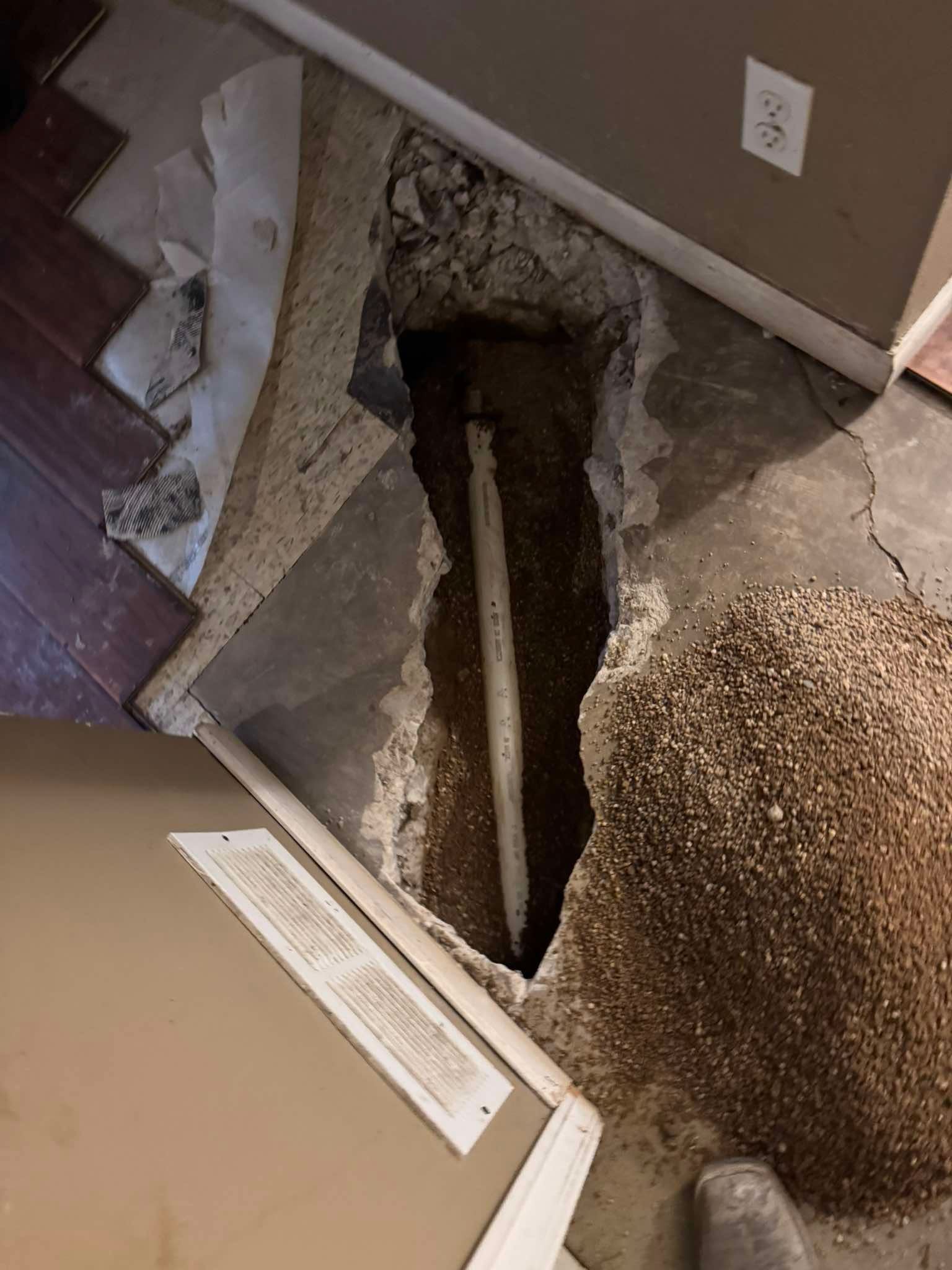 A trench cut into a concrete floor reveals a white pipe running through soil, with a pile of debris nearby.