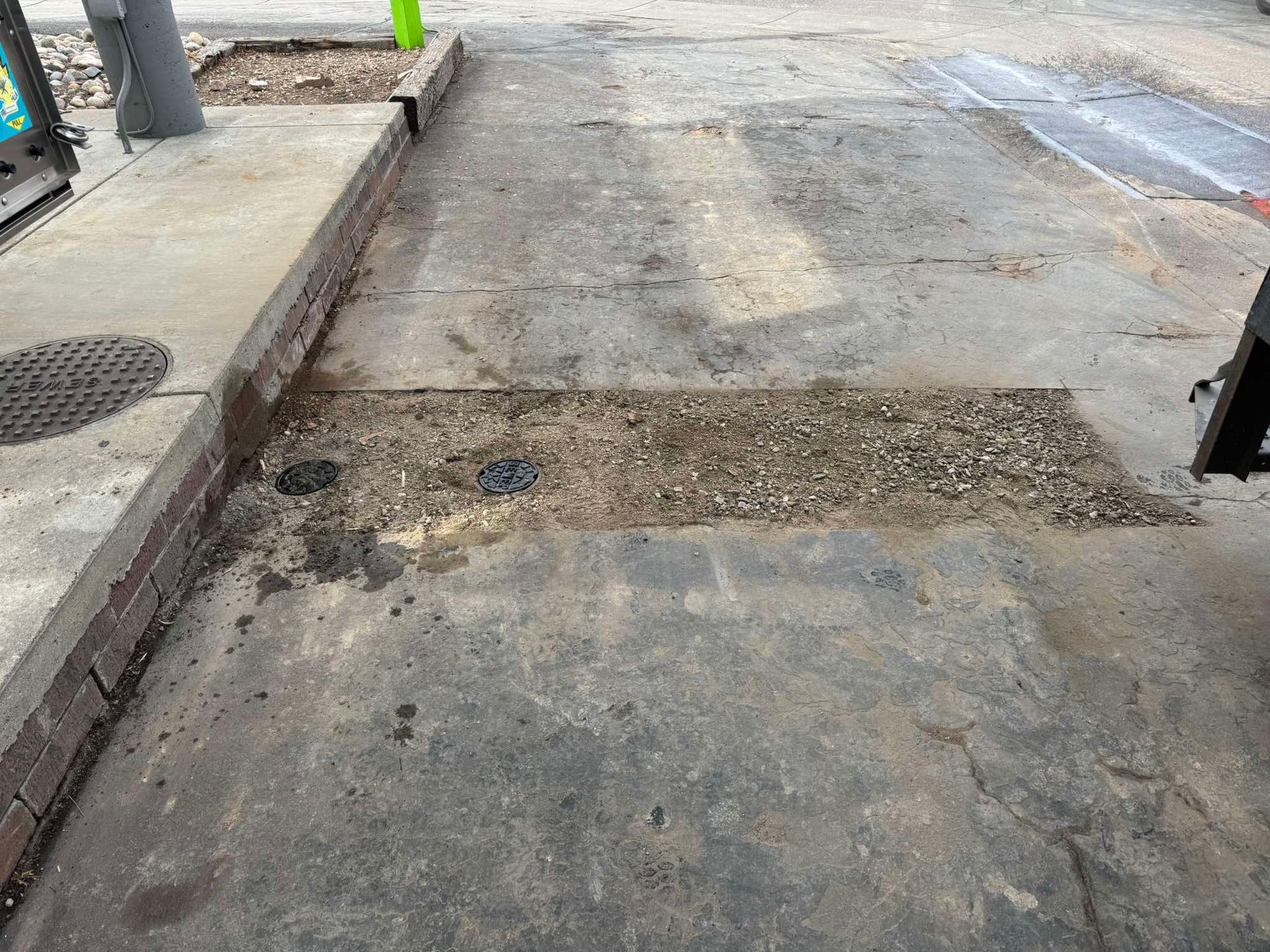 A concrete sidewalk featuring a damaged, rectangular patch filled with loose gravel and two utility access covers.