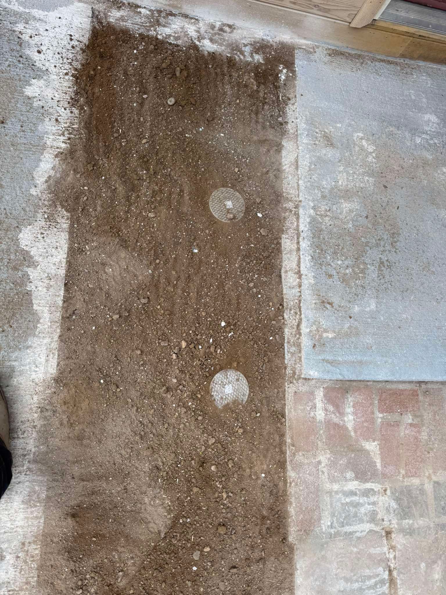 A rectangular strip of brown soil with two circular indentations on a concrete surface next to paved ground.