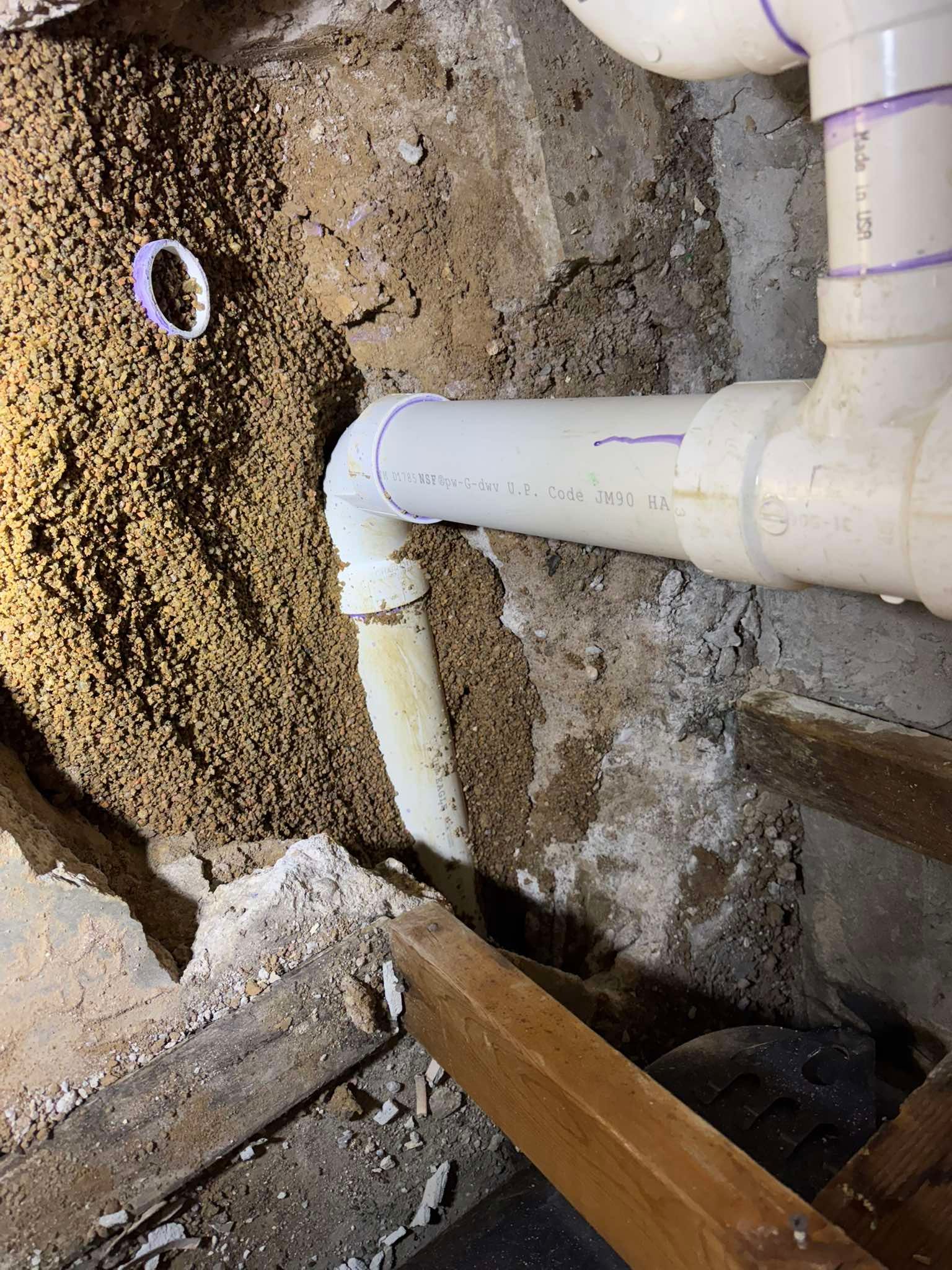White PVC plumbing pipes with purple primer are connected at a corner in a rough, dirt-filled crawlspace or wall cavity.