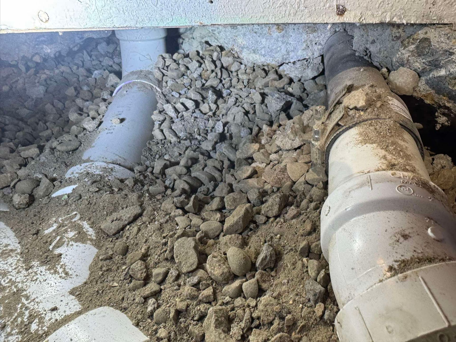 Two gray PVC pipes pass through a layer of dirt and gravel under a building foundation.