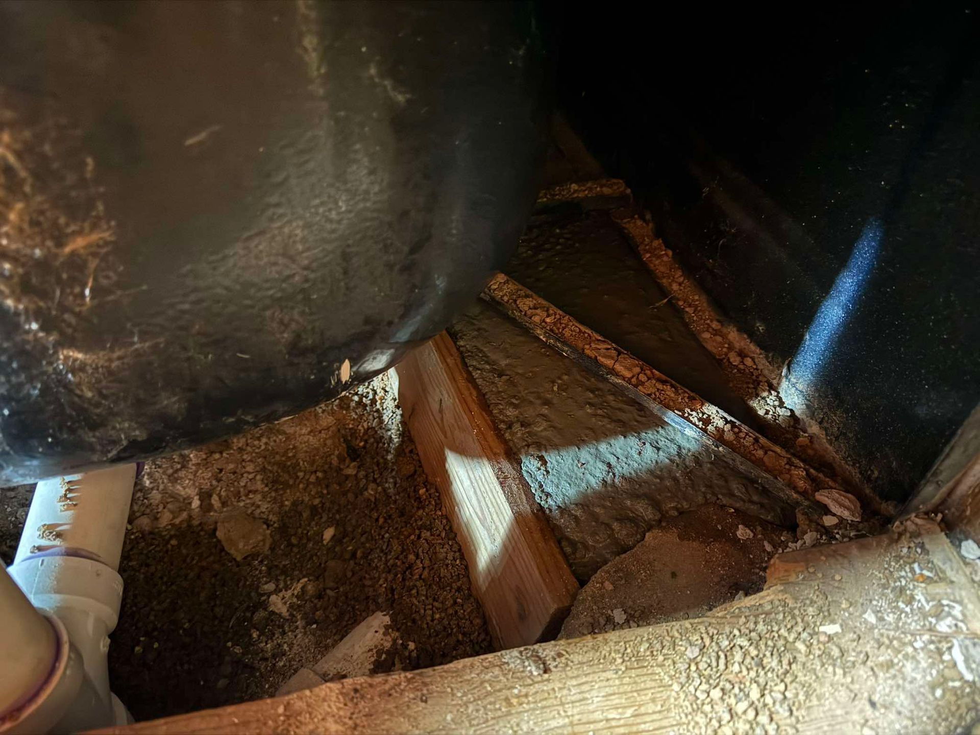 A dark, cluttered crawl space featuring wooden beams, exposed pipes, and loose debris beneath a large black object.
