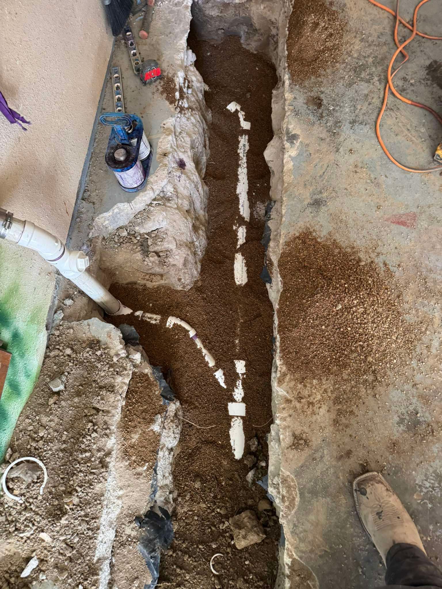 White PVC plumbing pipes are laid in a narrow dirt trench excavated through a concrete floor.