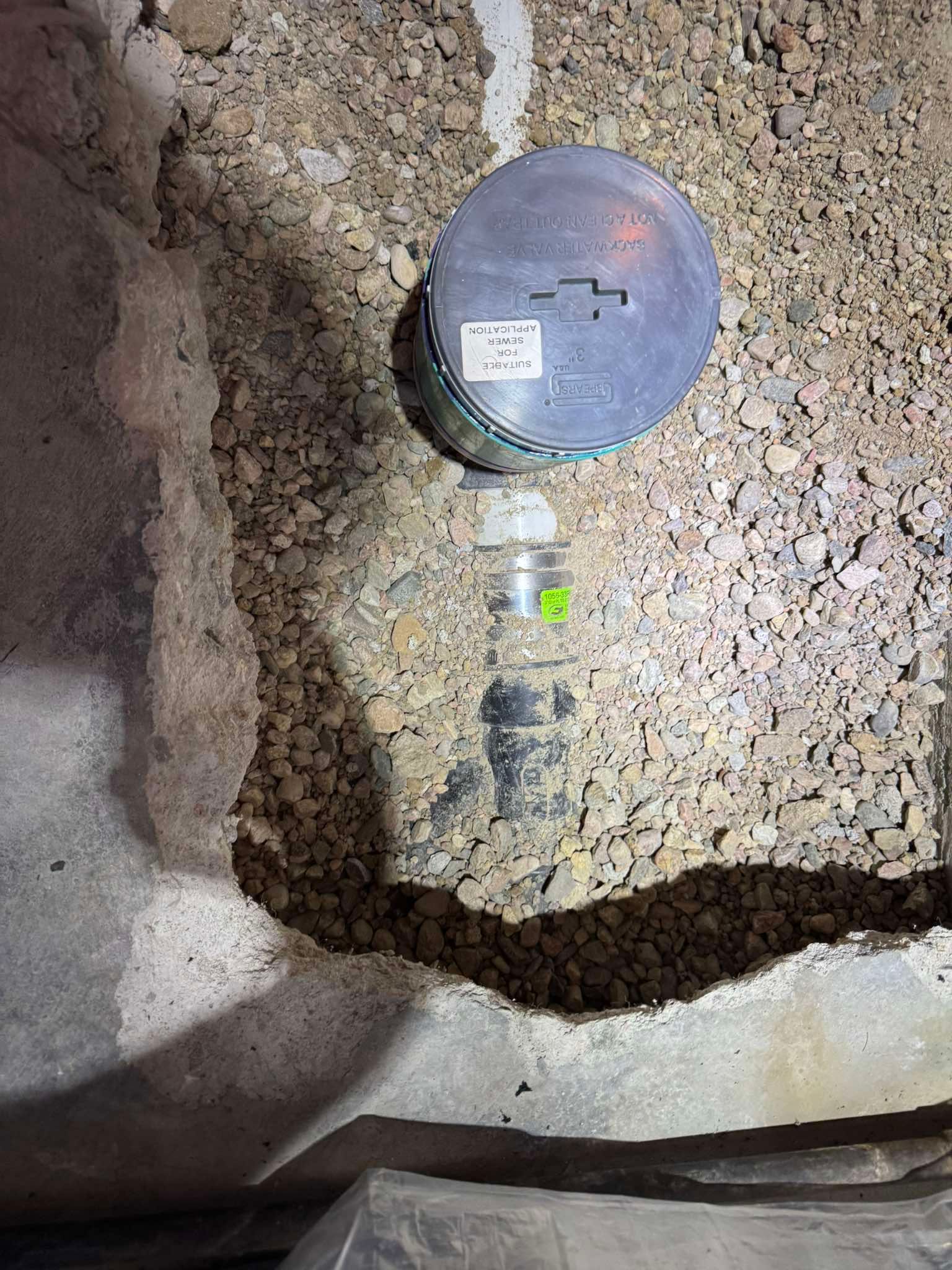 A circular metal junction box sits on a gravel bed next to a trench cut into a concrete floor.