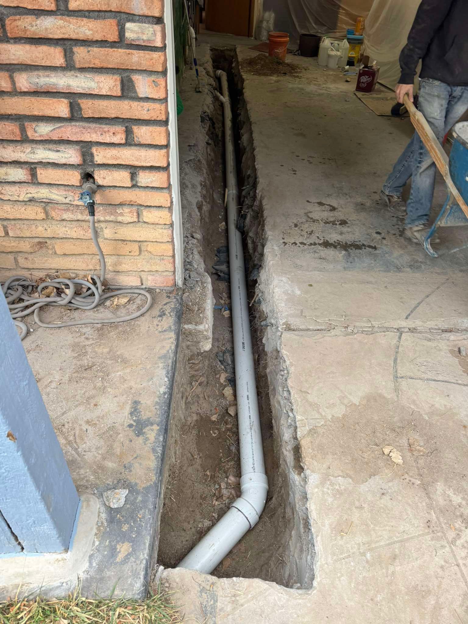 A grey PVC pipe lies in a narrow trench cut through a concrete patio next to a brick wall.