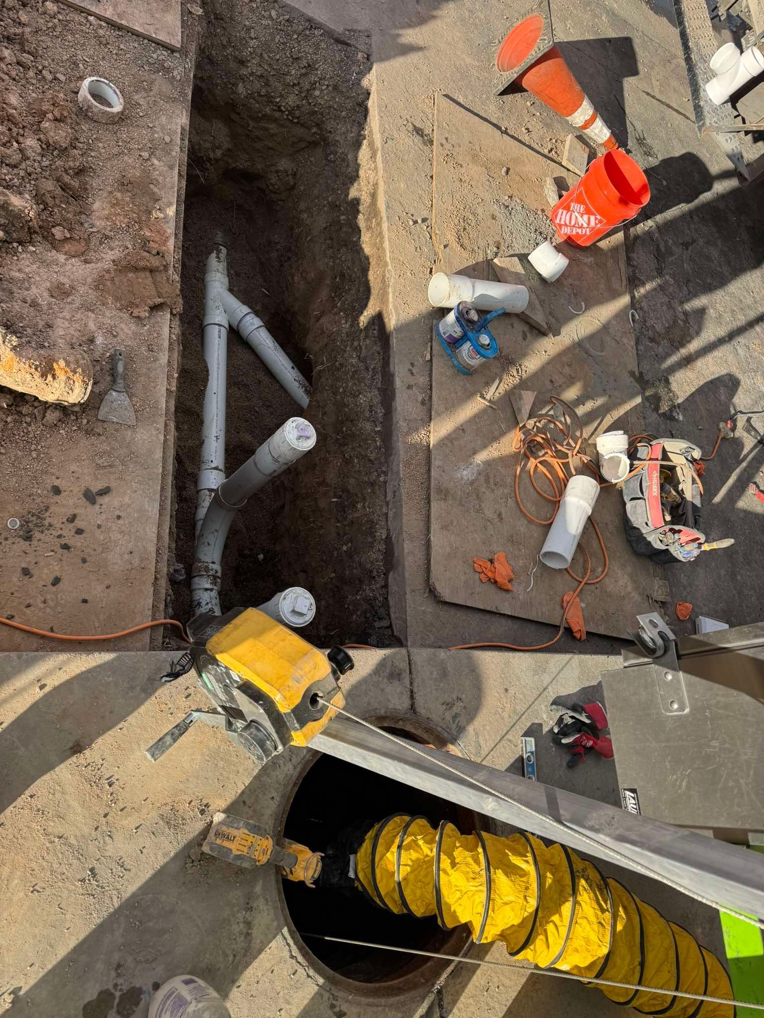 A construction trench with gray pipes, a yellow blower duct near a manhole, and various tools on the surrounding pavement.