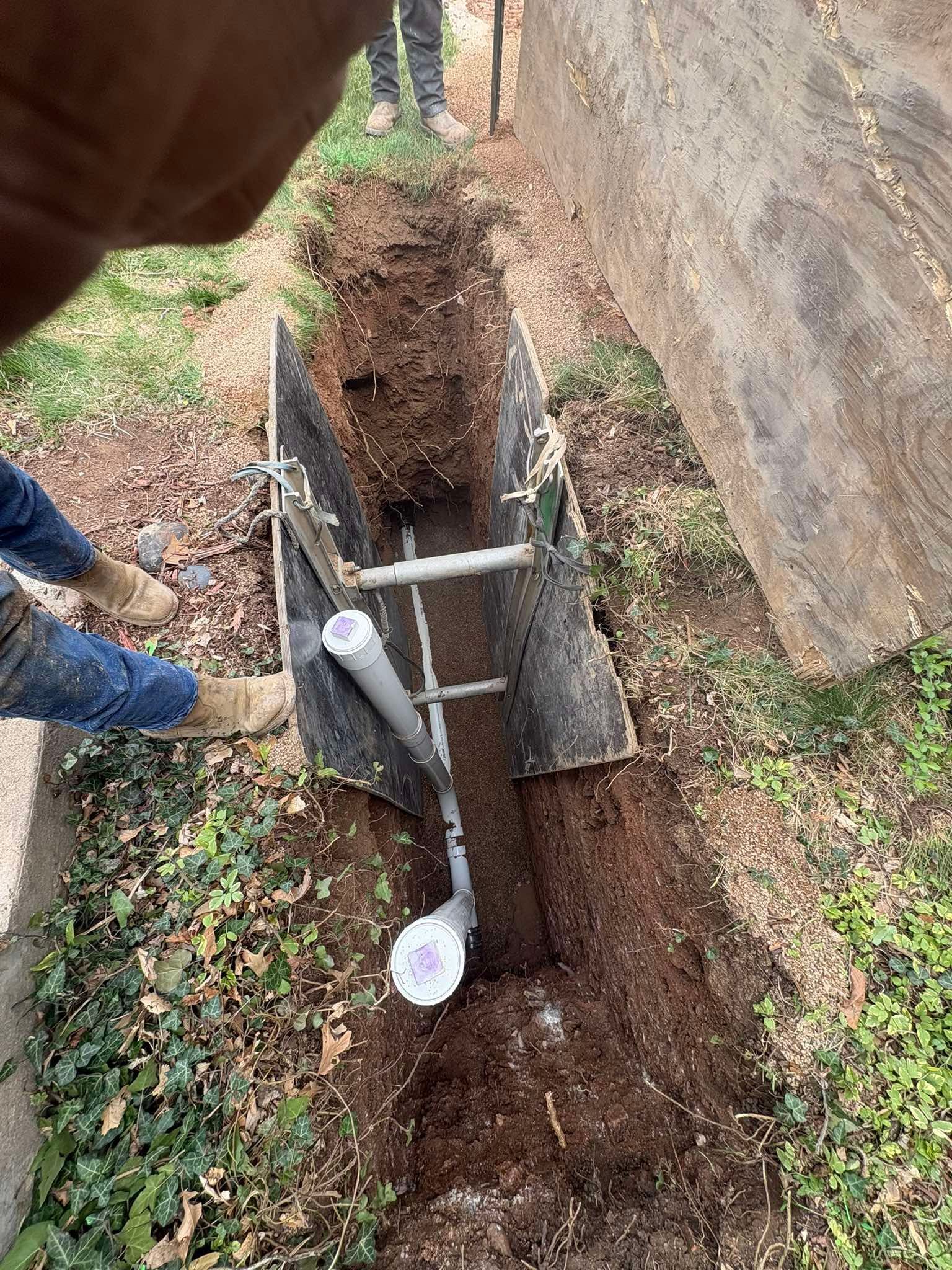 A person stands near a deep narrow trench in the dirt, shored up with metal plates and a PVC pipe installation inside.