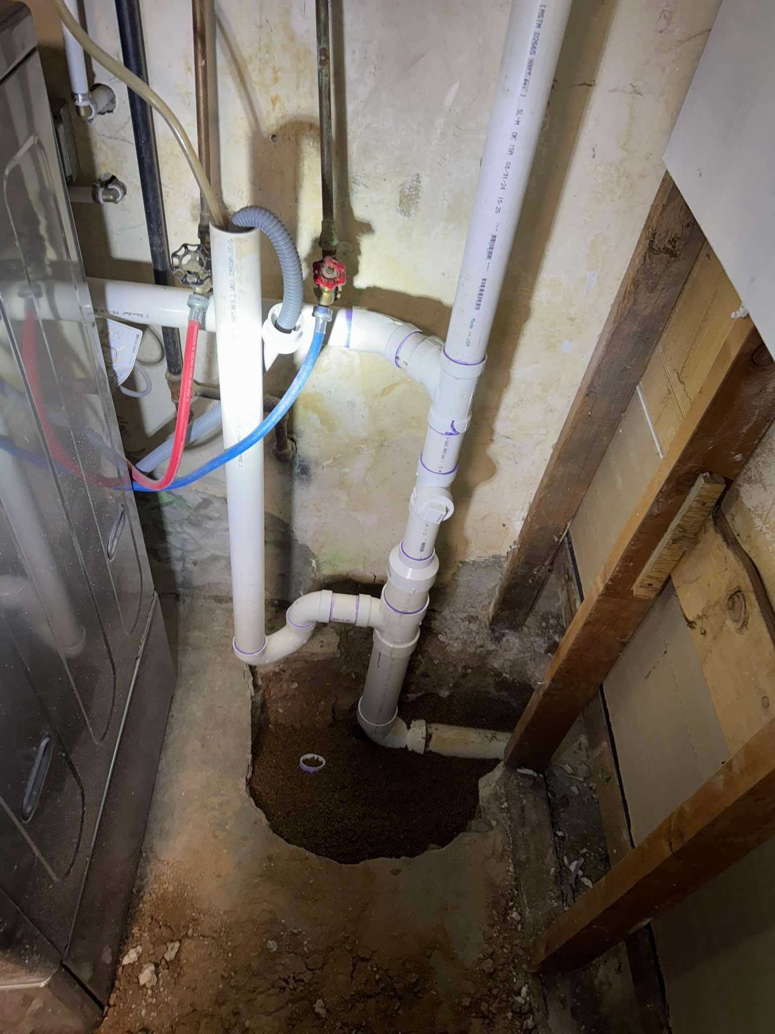 White PVC plumbing pipes, including a drain and supply lines, installed against a wall over a hole in a concrete floor.