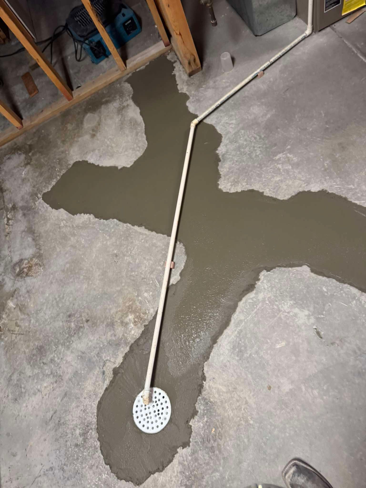 A white PVC pipe runs across a basement concrete floor to a circular floor drain set in wet, gray mortar.