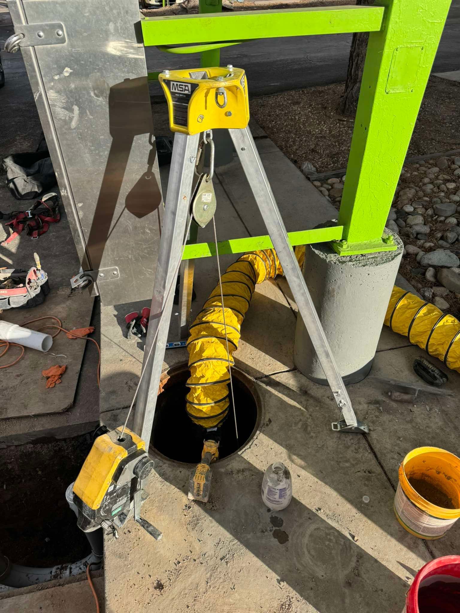 A tripod with a winch stands over an open manhole, with a yellow ventilation duct leading inside.
