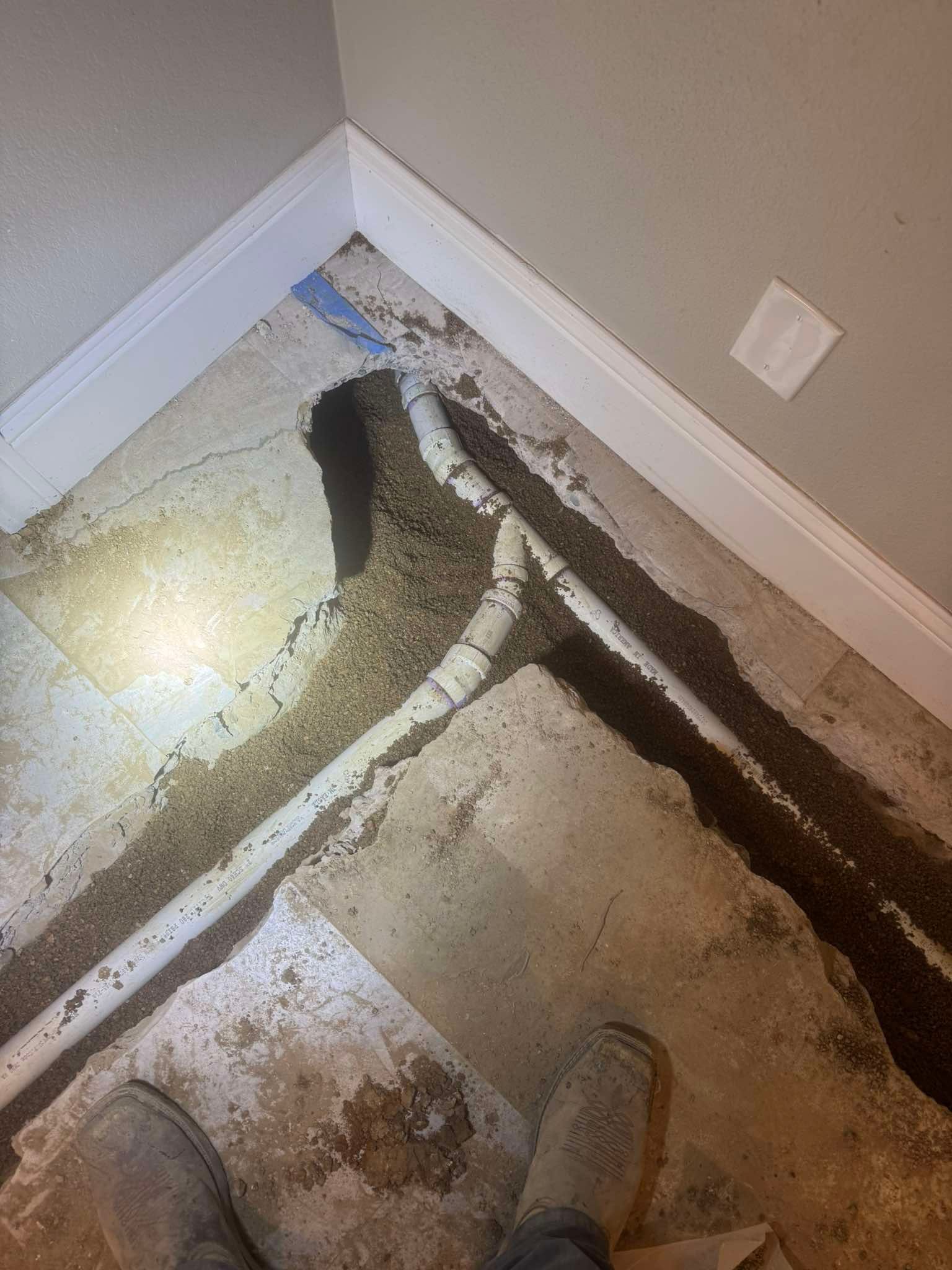 A view looking down at a white PVC pipe exposed in a trench cut through a concrete floor in a room corner.