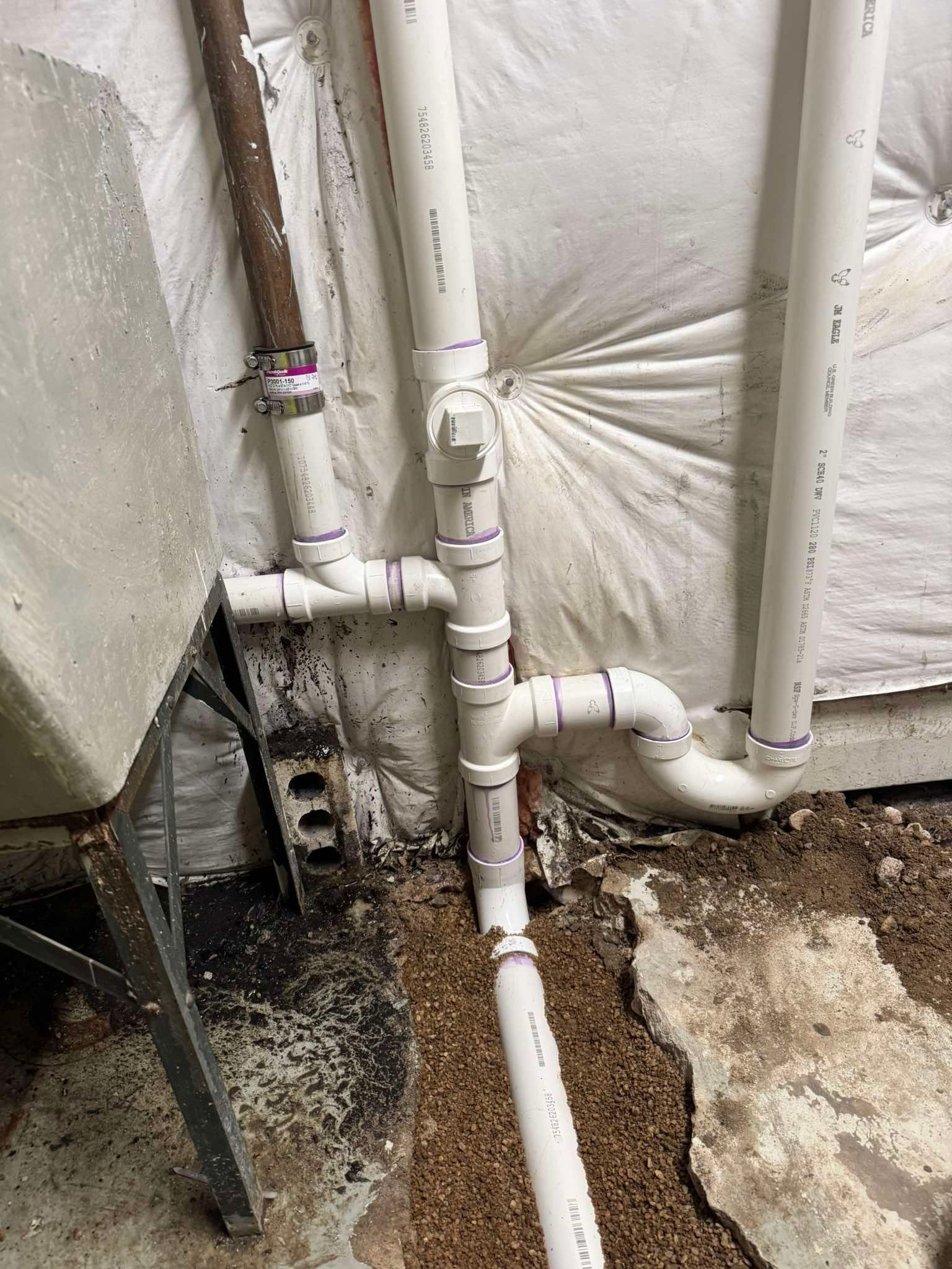 A white PVC pipe plumbing assembly connecting a drain line to a vertical main stack against an insulated basement wall.