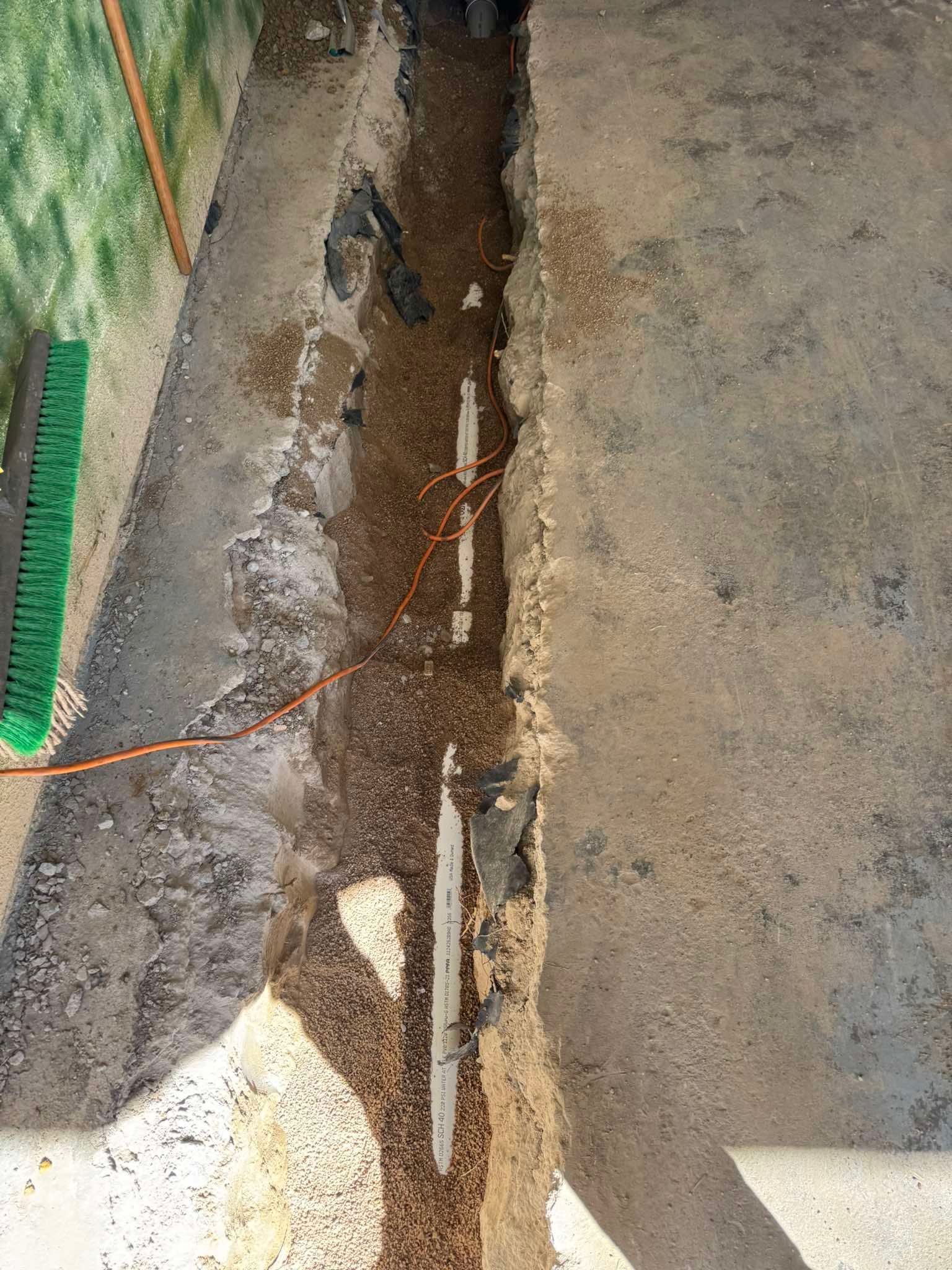A narrow trench cut into a concrete floor, revealing dirt, gravel, and an exposed white pipe running down the center.