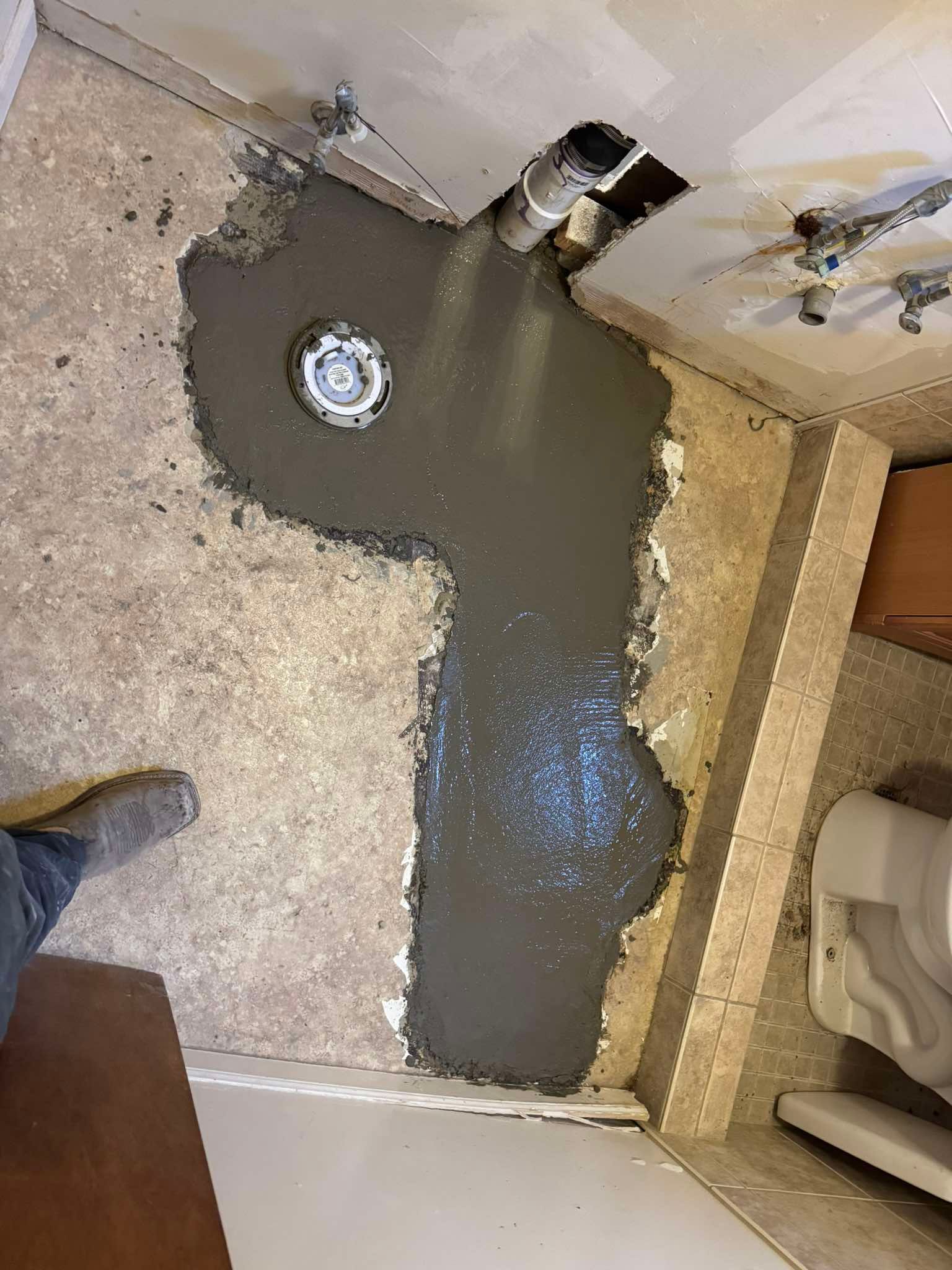 A bathroom floor renovation in progress with a newly poured gray concrete patch surrounding a floor drain and pipe.