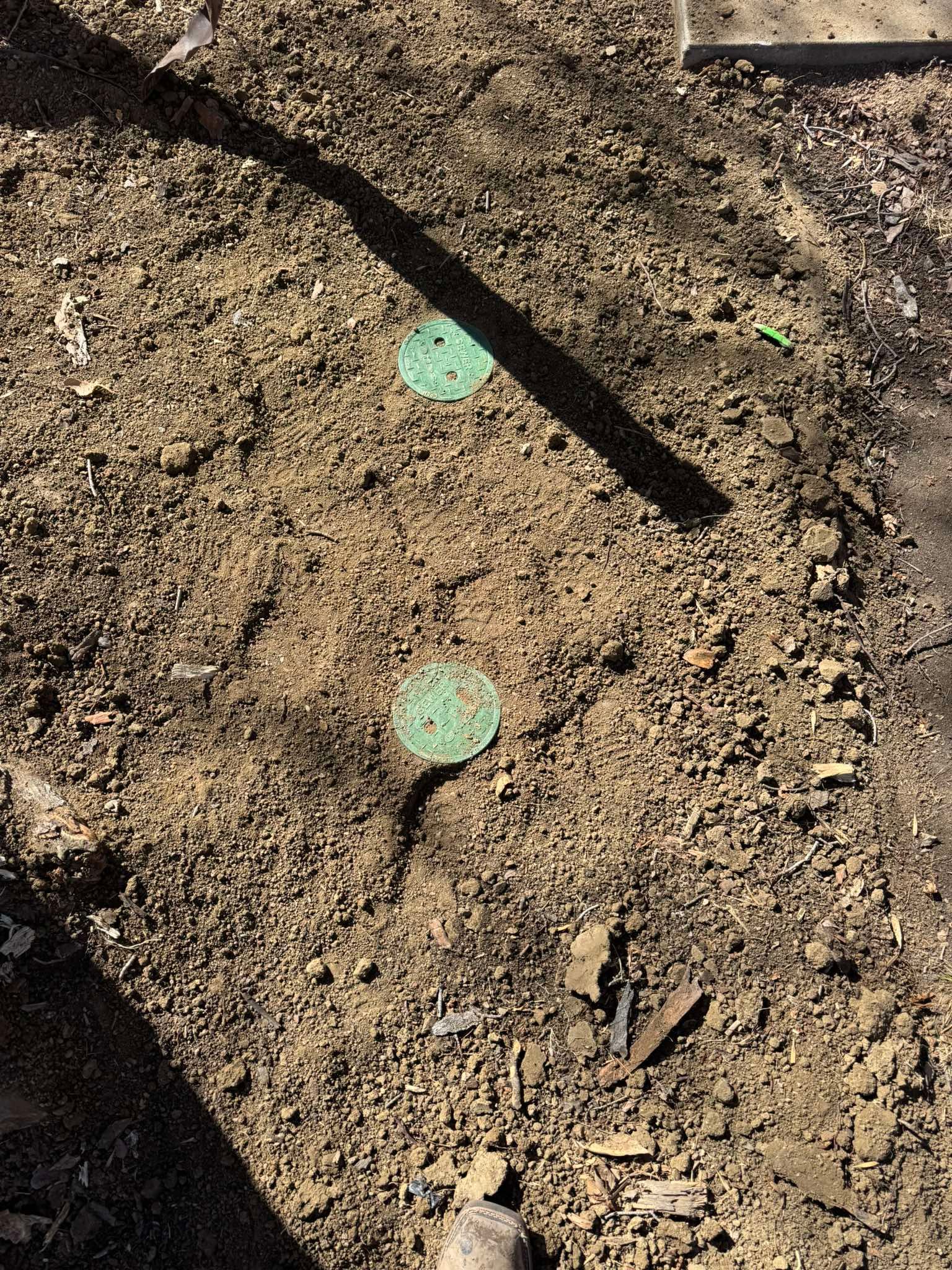 Two small, round, green irrigation valve box covers embedded in dry, dirt-covered soil.
