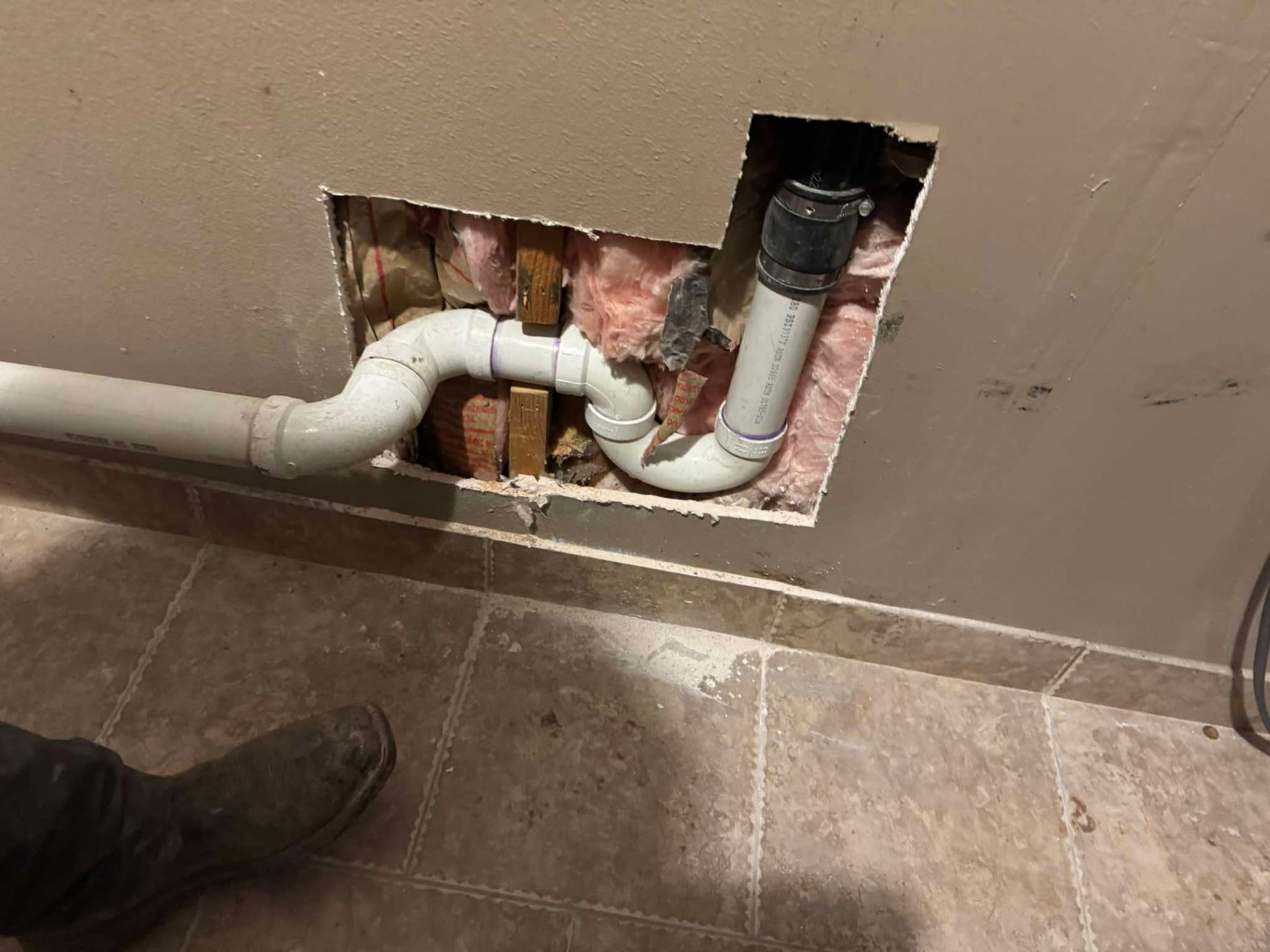 A section of white PVC plumbing pipe with an S-curve configuration installed inside an open hole in a residential wall.