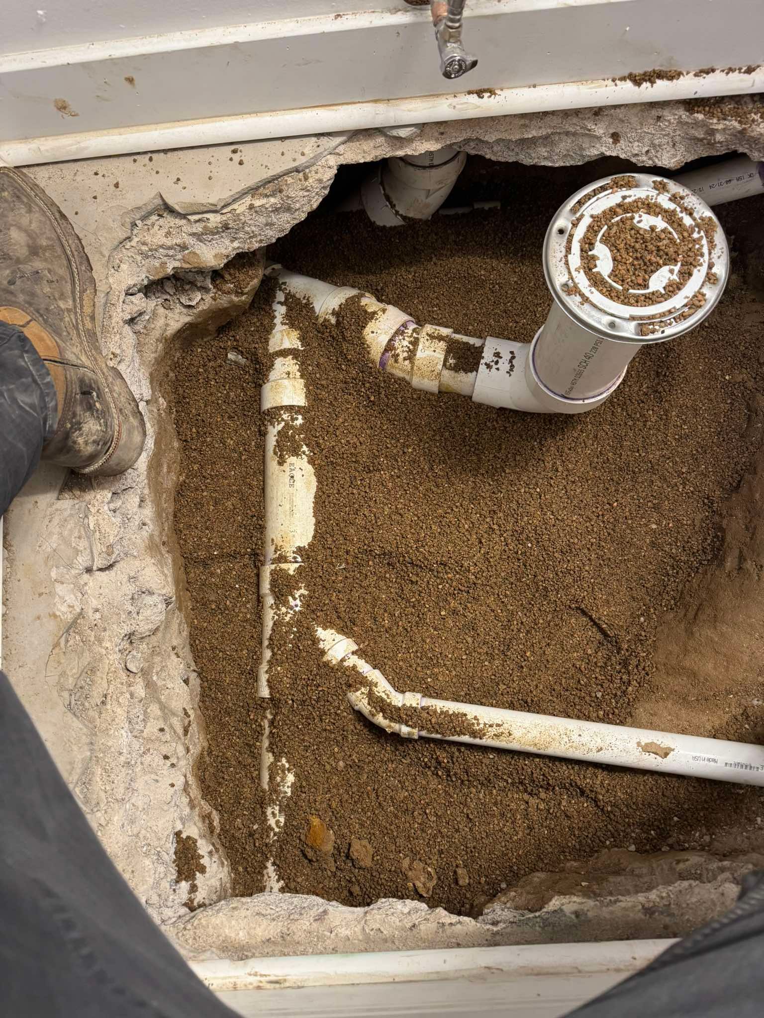 A hole cut into a concrete floor shows white PVC plumbing pipes partially embedded in brown, loose soil.