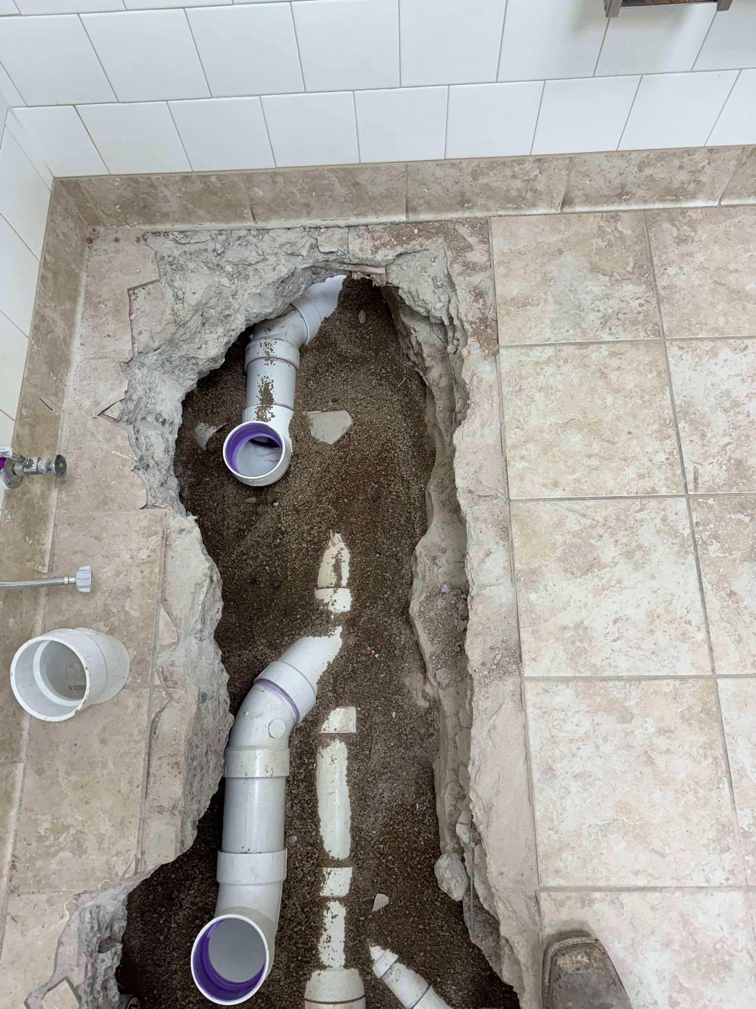 A trench cut into a tiled floor exposes white PVC plumbing pipes set in gravel, indicating an active bathroom renovation.