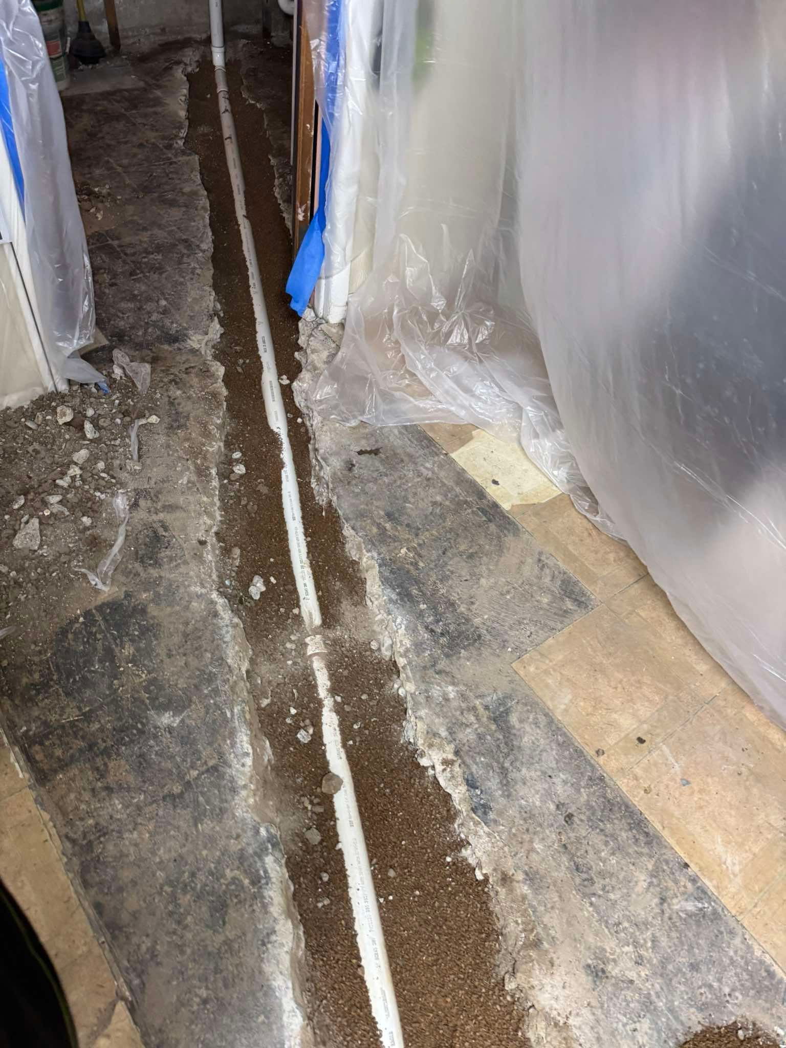 A white PVC pipe runs through a narrow, dirt-filled trench cut into a concrete floor, framed by plastic sheeting.