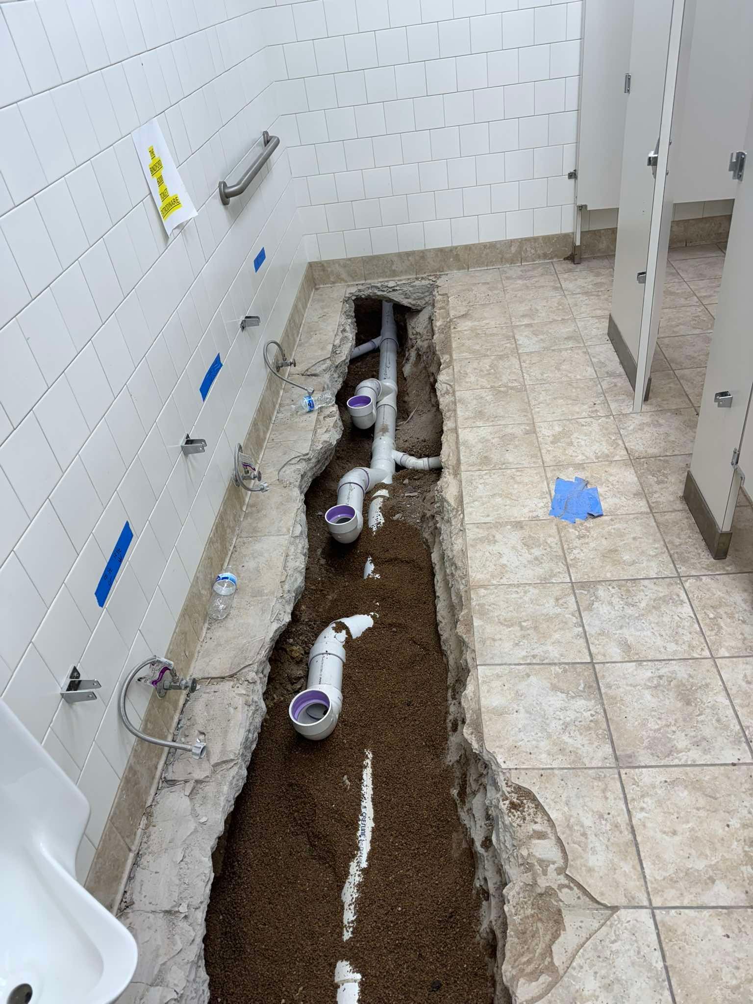 A partially renovated restroom floor with a trench cut into the tile, exposing PVC plumbing pipes connected to urinals.