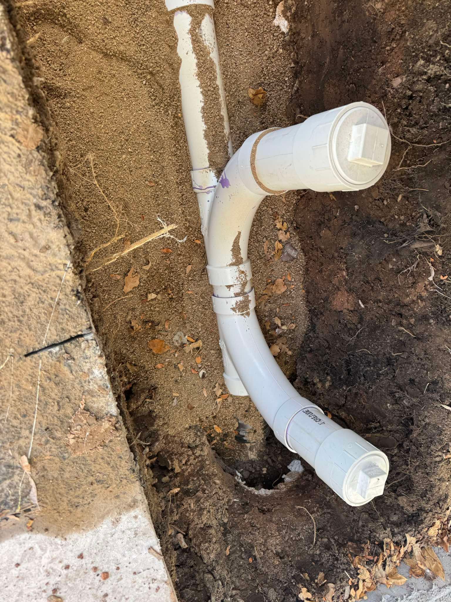 A PVC pipe assembly with two capped cleanout branches installed in a narrow, dirt-filled trench.