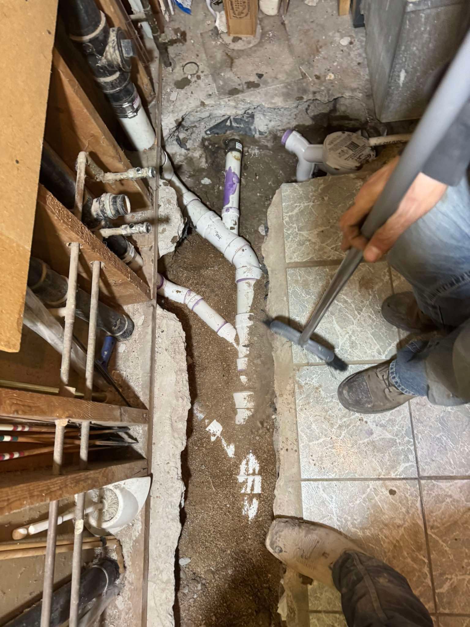 A worker cleans debris from a trench in a concrete floor containing newly installed white PVC plumbing pipes.