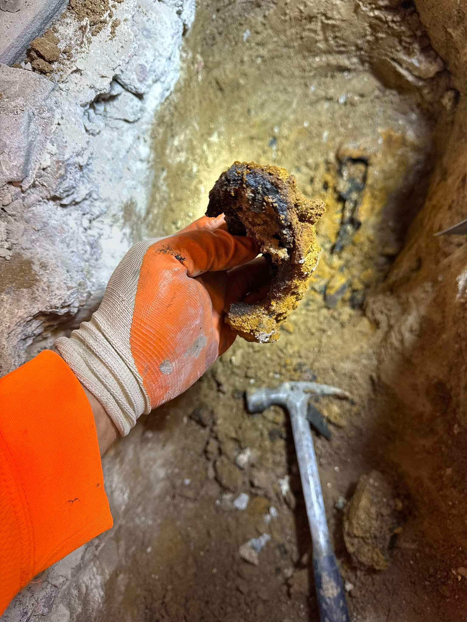 A gloved hand holds a piece of corroded metal in a hole in the ground, with a hammer lying in the dirt nearby.