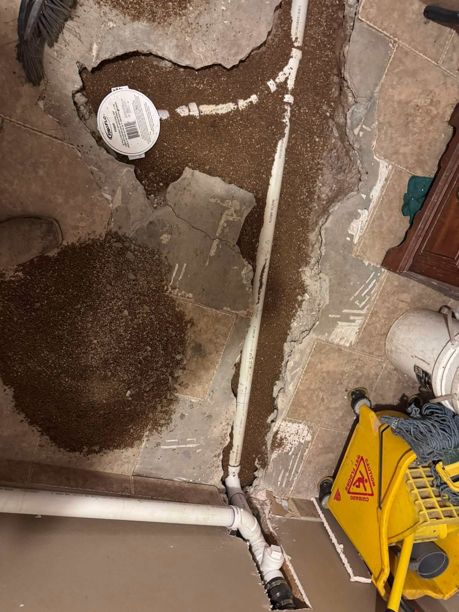 A trench cut into a concrete floor, revealing white plumbing pipes surrounded by gravel, with a yellow mop bucket nearby.