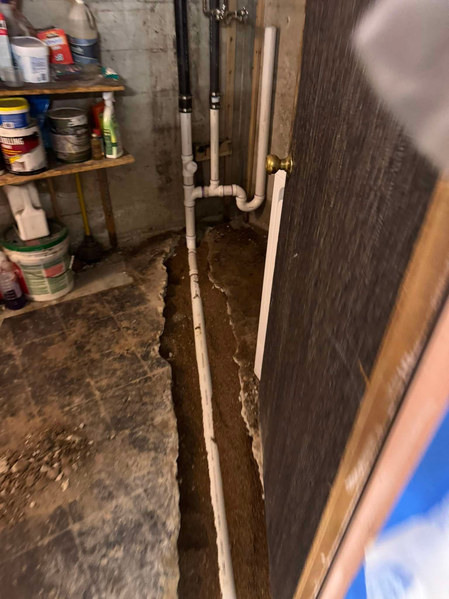 A basement floor with a trench cut into the concrete revealing a white plastic pipe running along the dirt underneath.