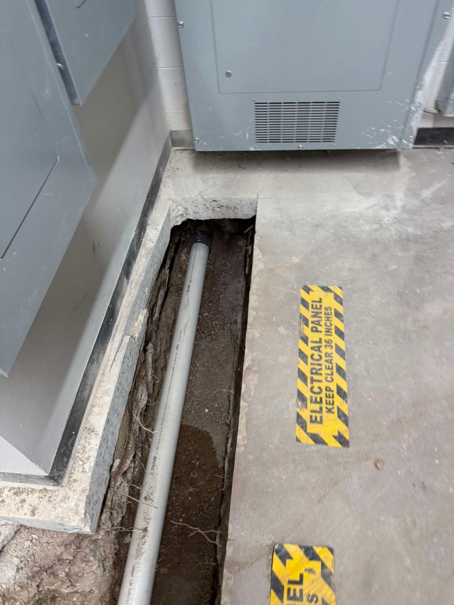 A concrete floor with a rectangular trench exposing a PVC pipe below, next to yellow electrical panel warning markers.