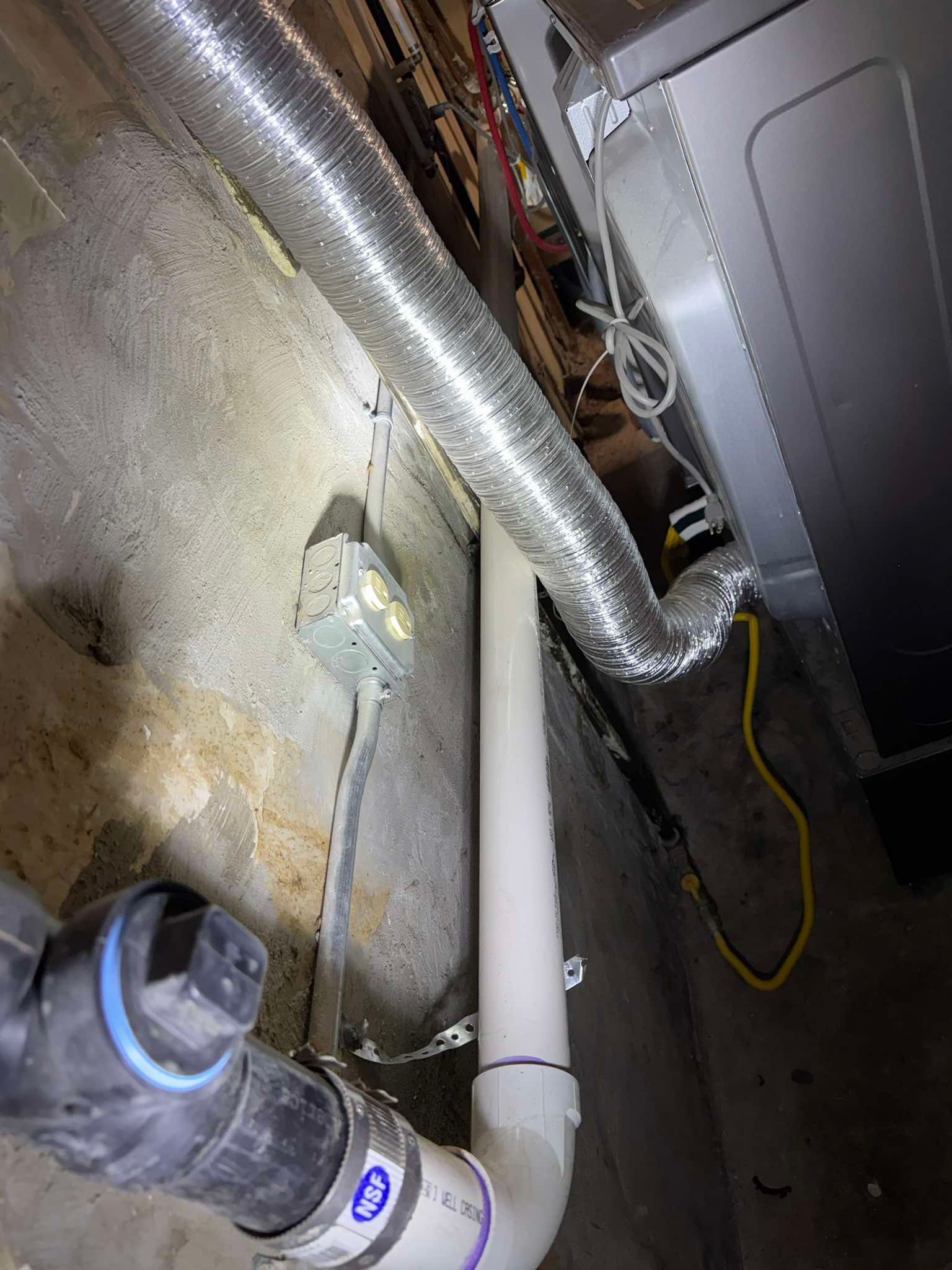 A utility room corner showing a white PVC pipe, silver dryer vent tubing, and an electrical outlet on a concrete wall.