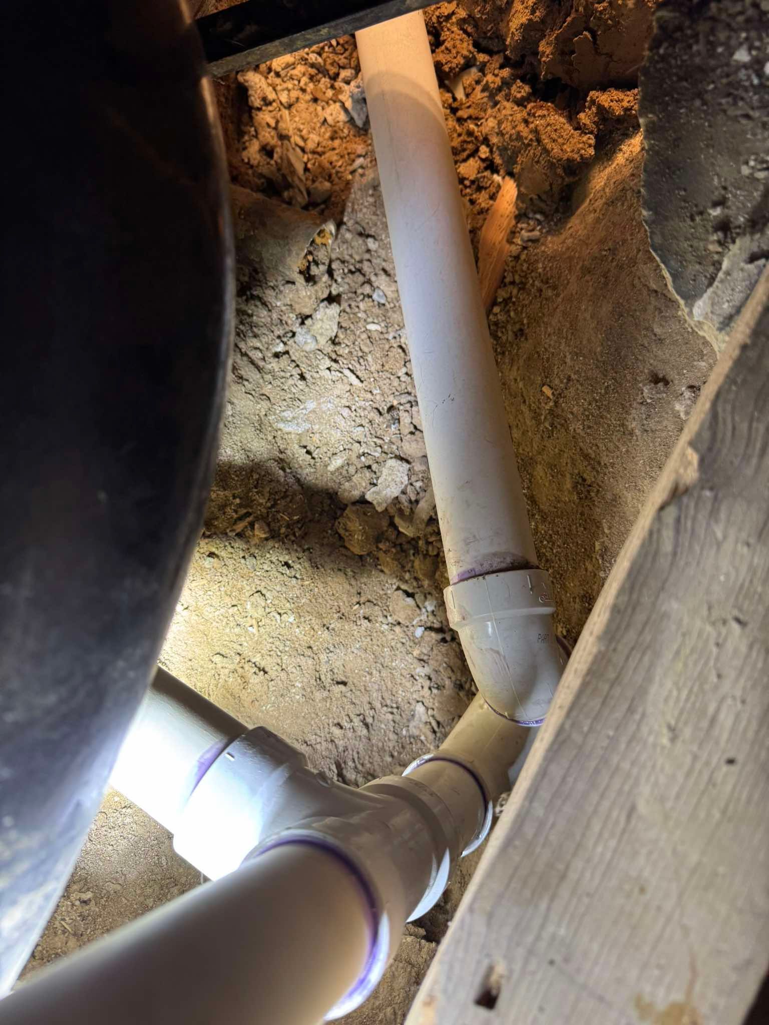 A close-up view of white PVC plumbing pipes connected with a T-fitting, set within a crawl space or wall cavity.