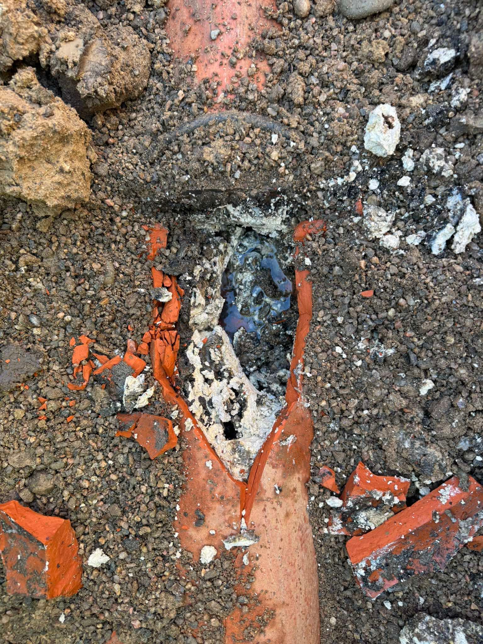 A broken orange clay pipe section buried in soil, revealing dark debris inside the damaged area.
