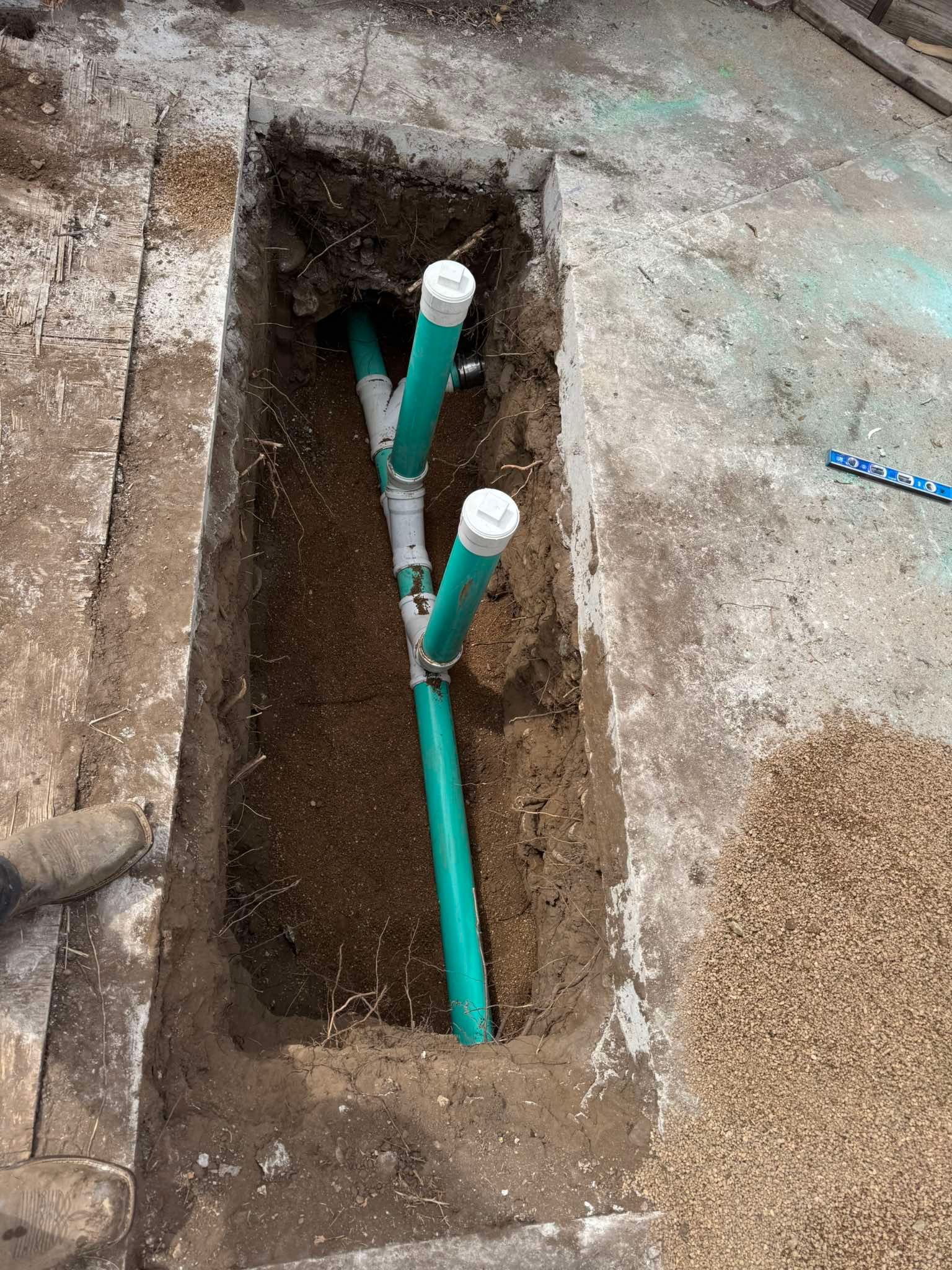 Green and white PVC plumbing pipes installed in a narrow, rectangular dirt trench cut into a concrete floor.