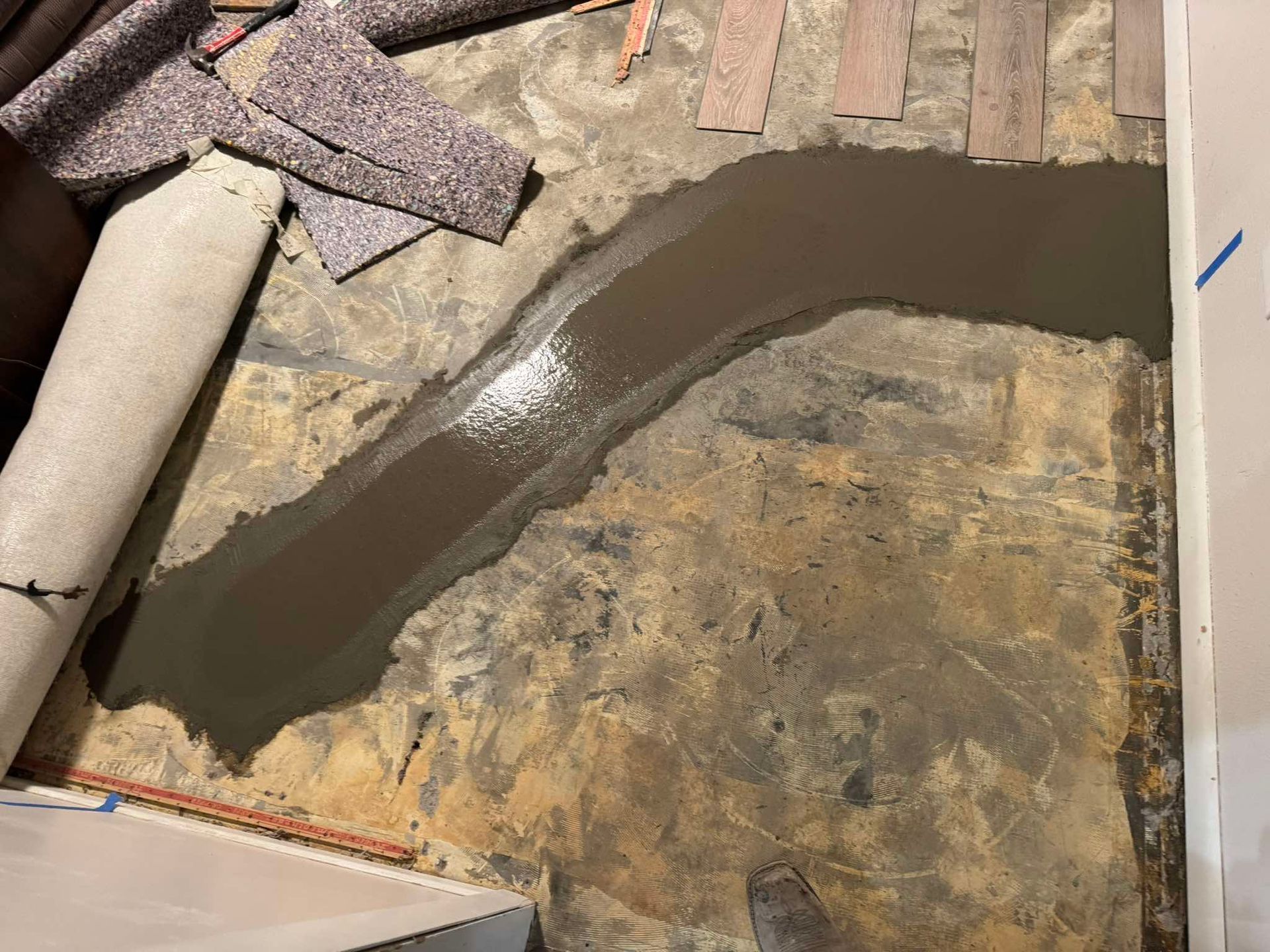 A trench in a concrete floor filled with wet, dark-gray cement filler, next to a rolled-up piece of carpet.
