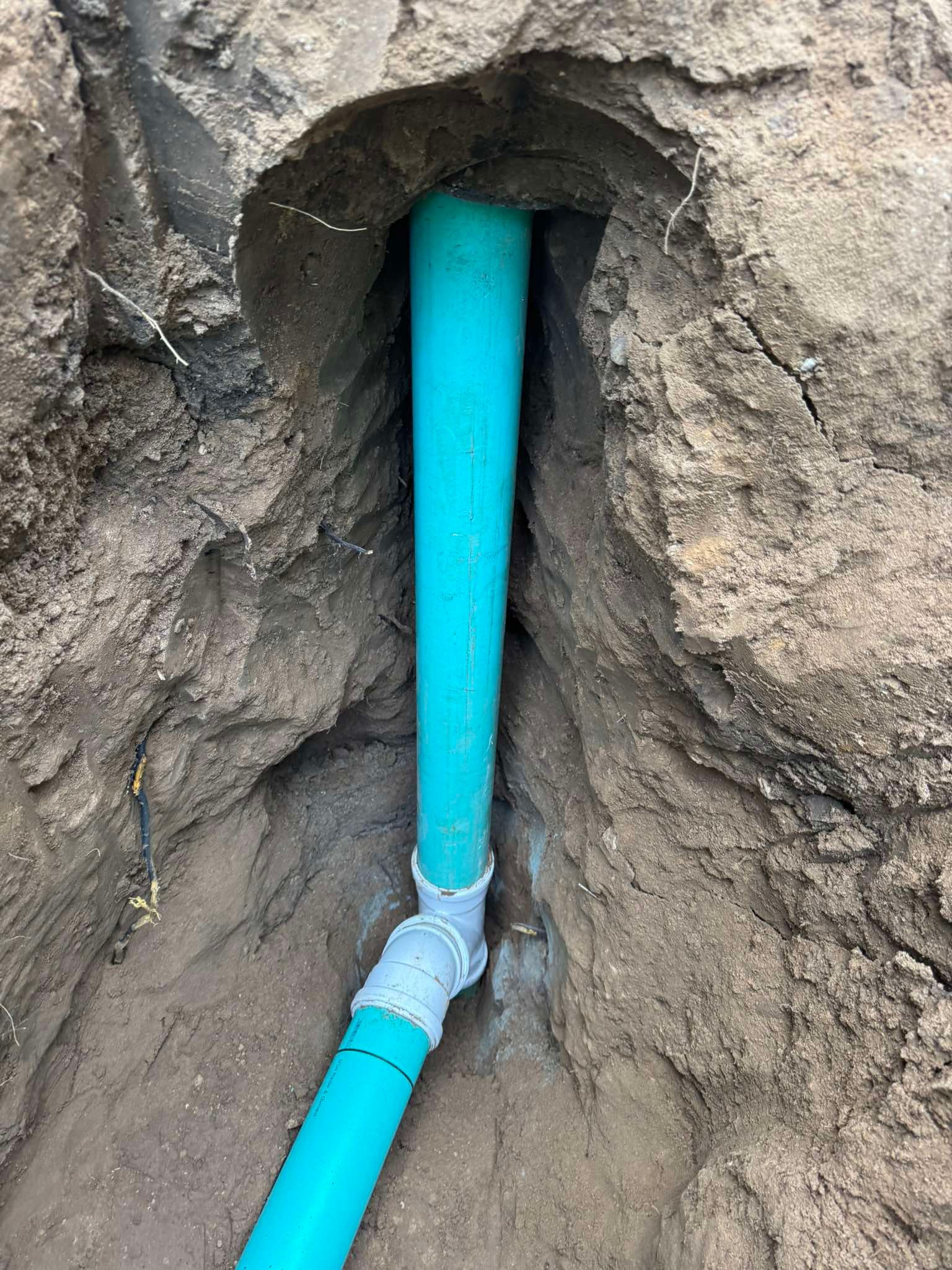 A green PVC pipe connection in a narrow dirt trench, showing a grey 90-degree elbow joint.