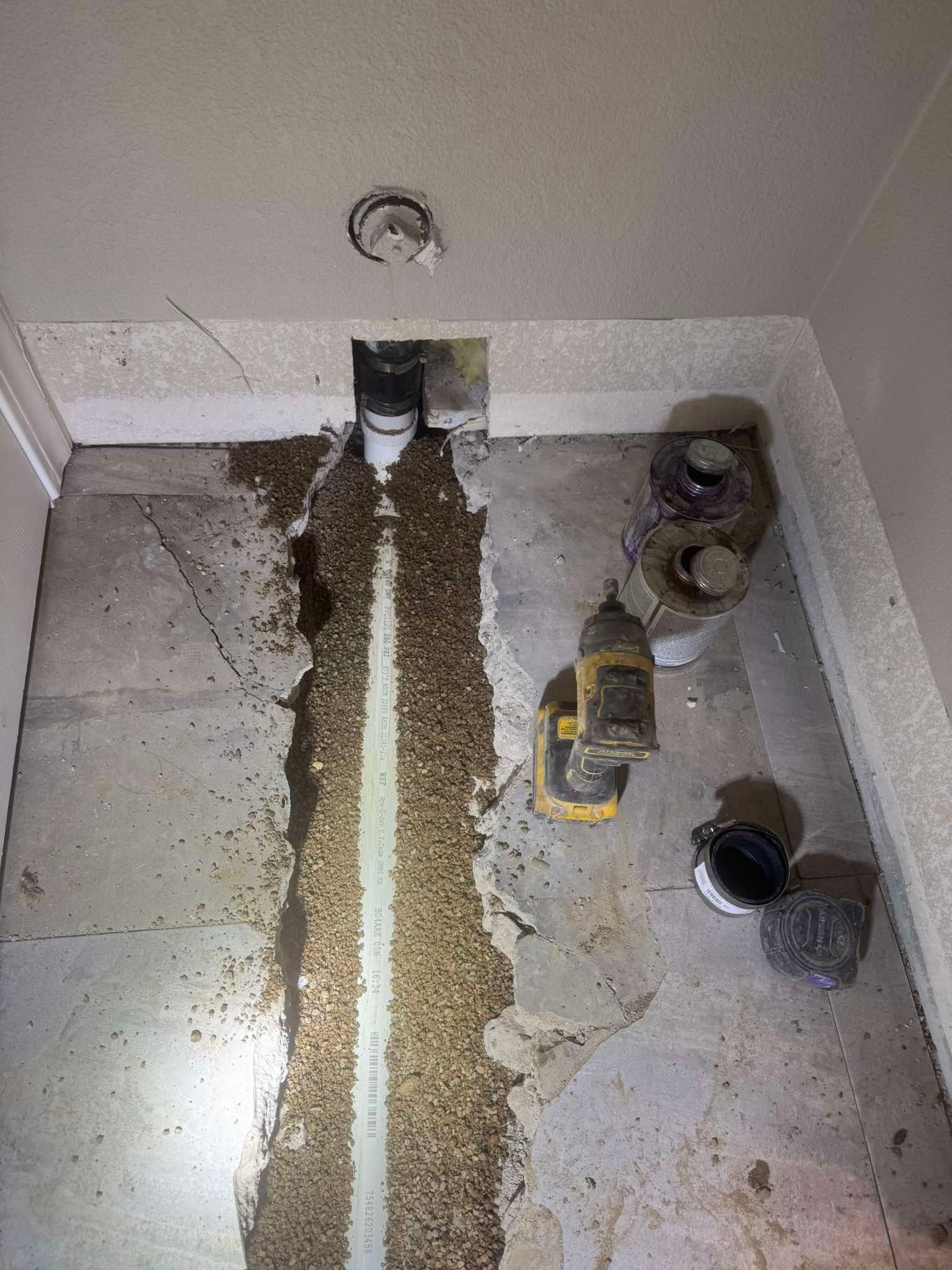An indoor concrete floor with a long, narrow trench dug out, exposing a new white pipe, with construction tools nearby.