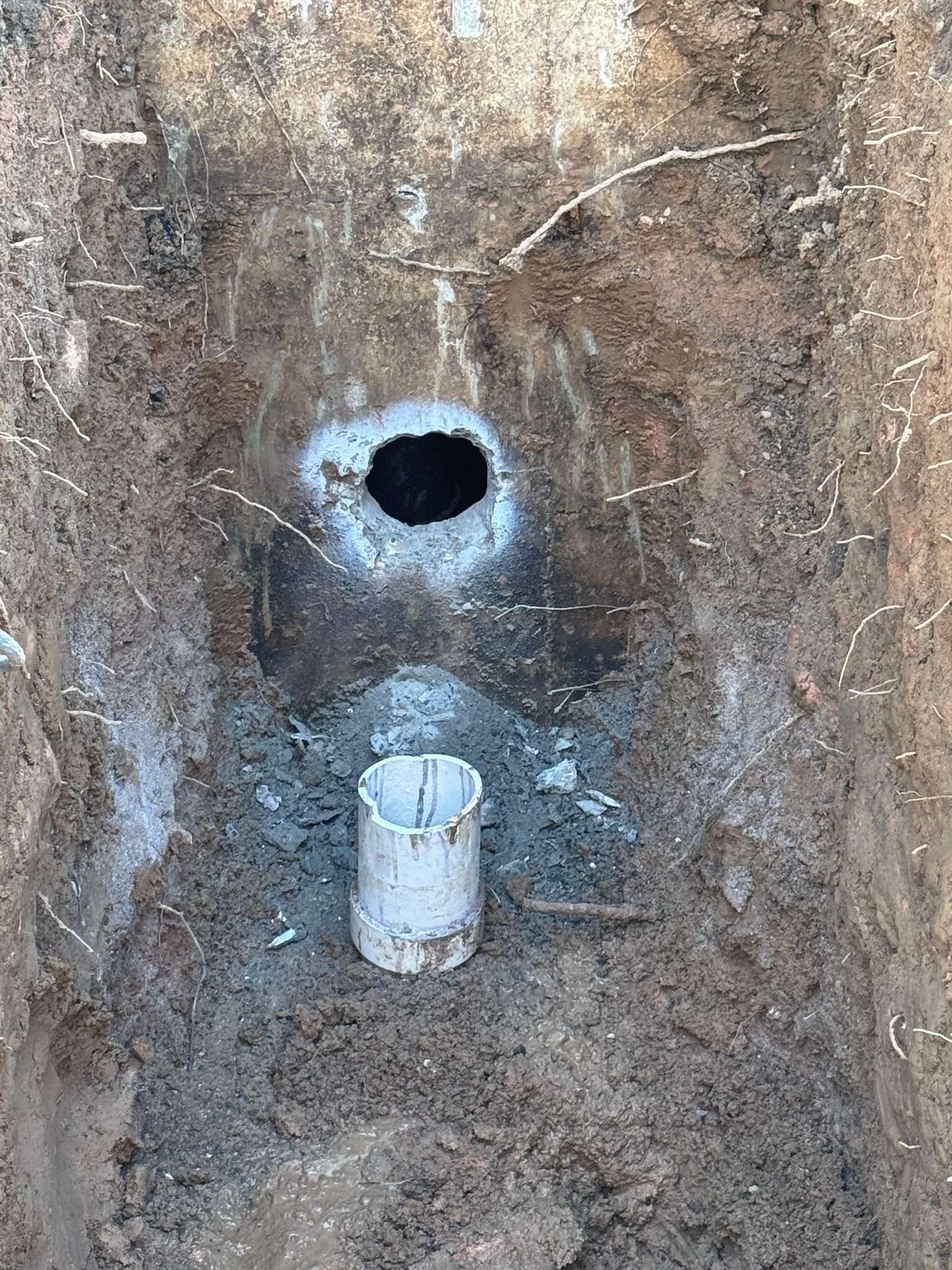 An excavated dirt pit shows a circular dark opening in the wall above a short, white plastic pipe protruding from the ground.