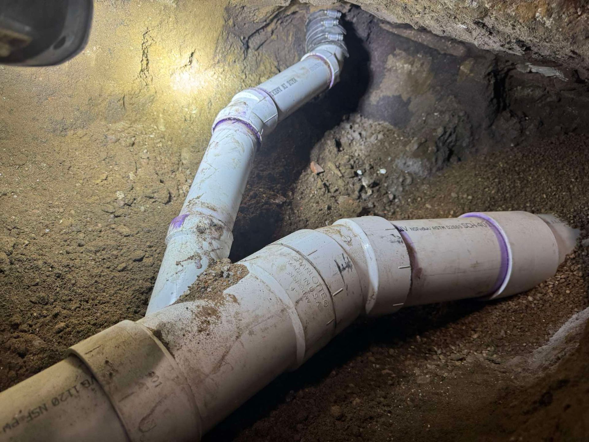 A newly installed white PVC pipe system consisting of a main line with a 45-degree angle branch, set in a dirt trench.