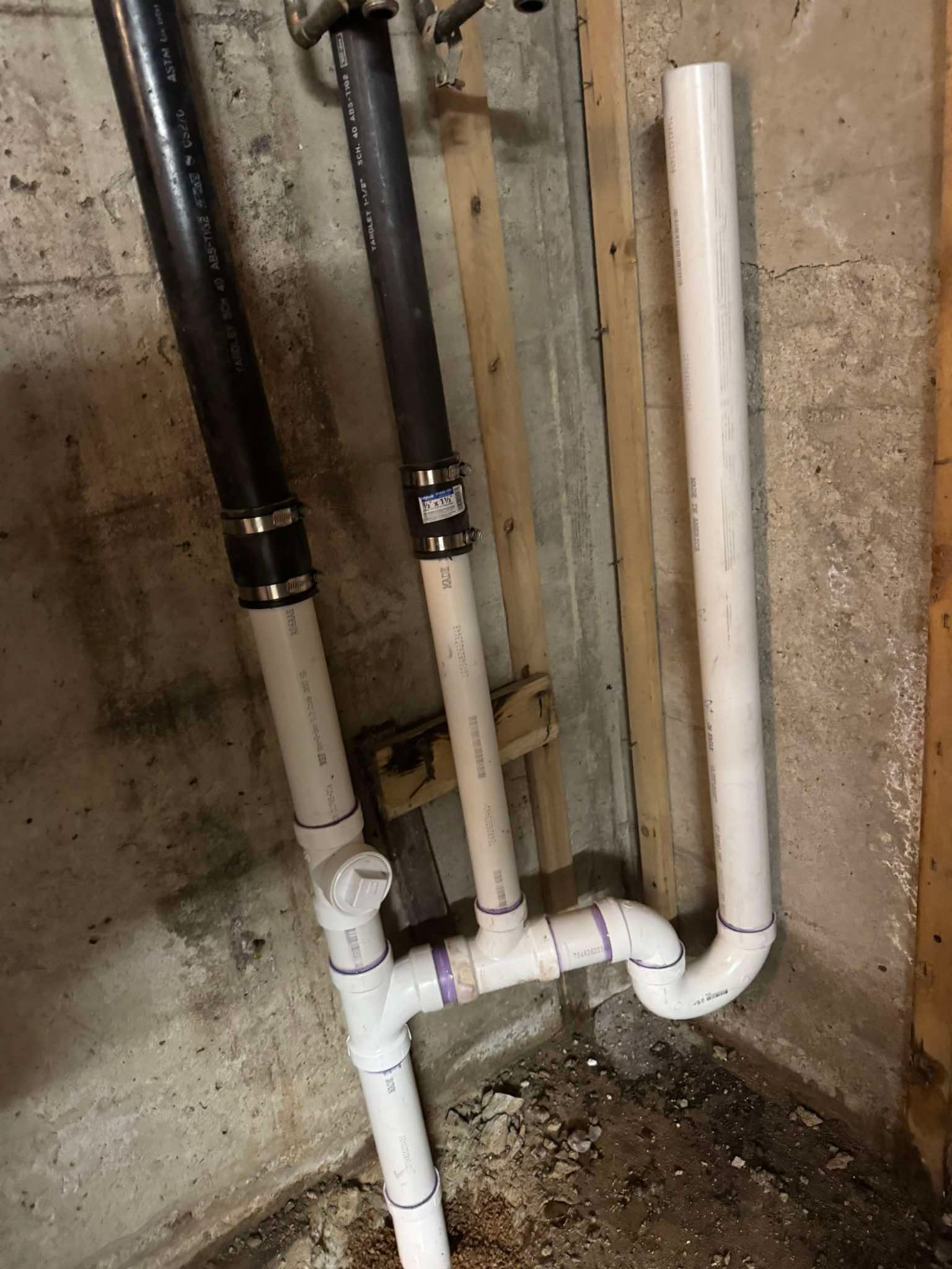 A vertical white PVC pipe assembly with two black pipe connections attached to a concrete wall.