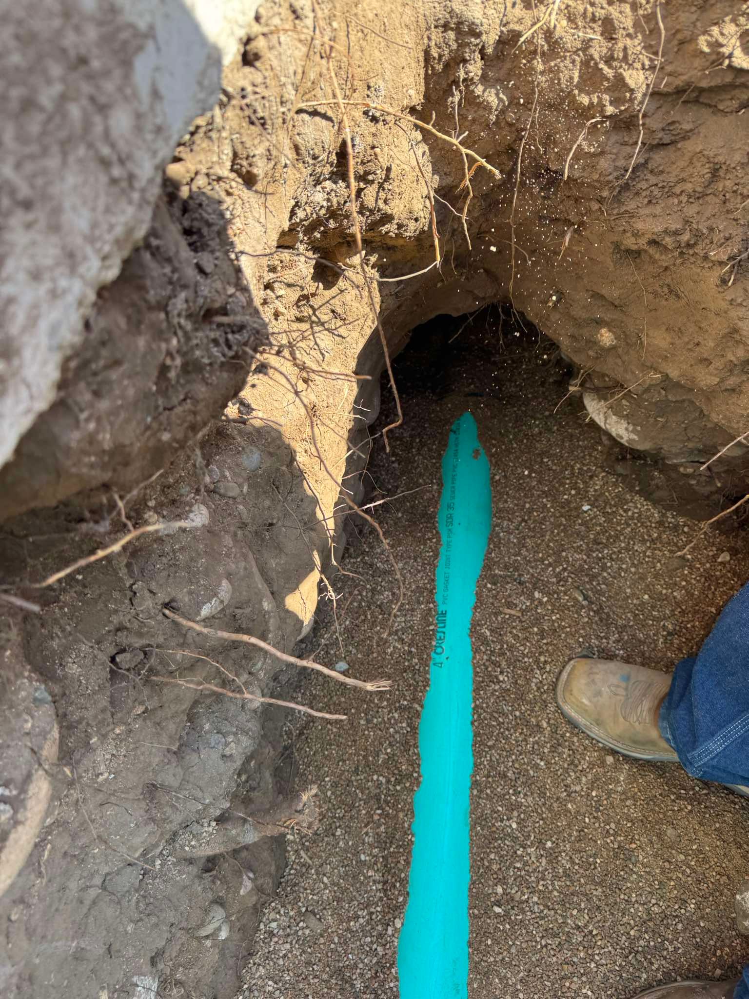 A blue pipe laid in a narrow dirt trench, viewed from above, with a person's boot visible in the corner.