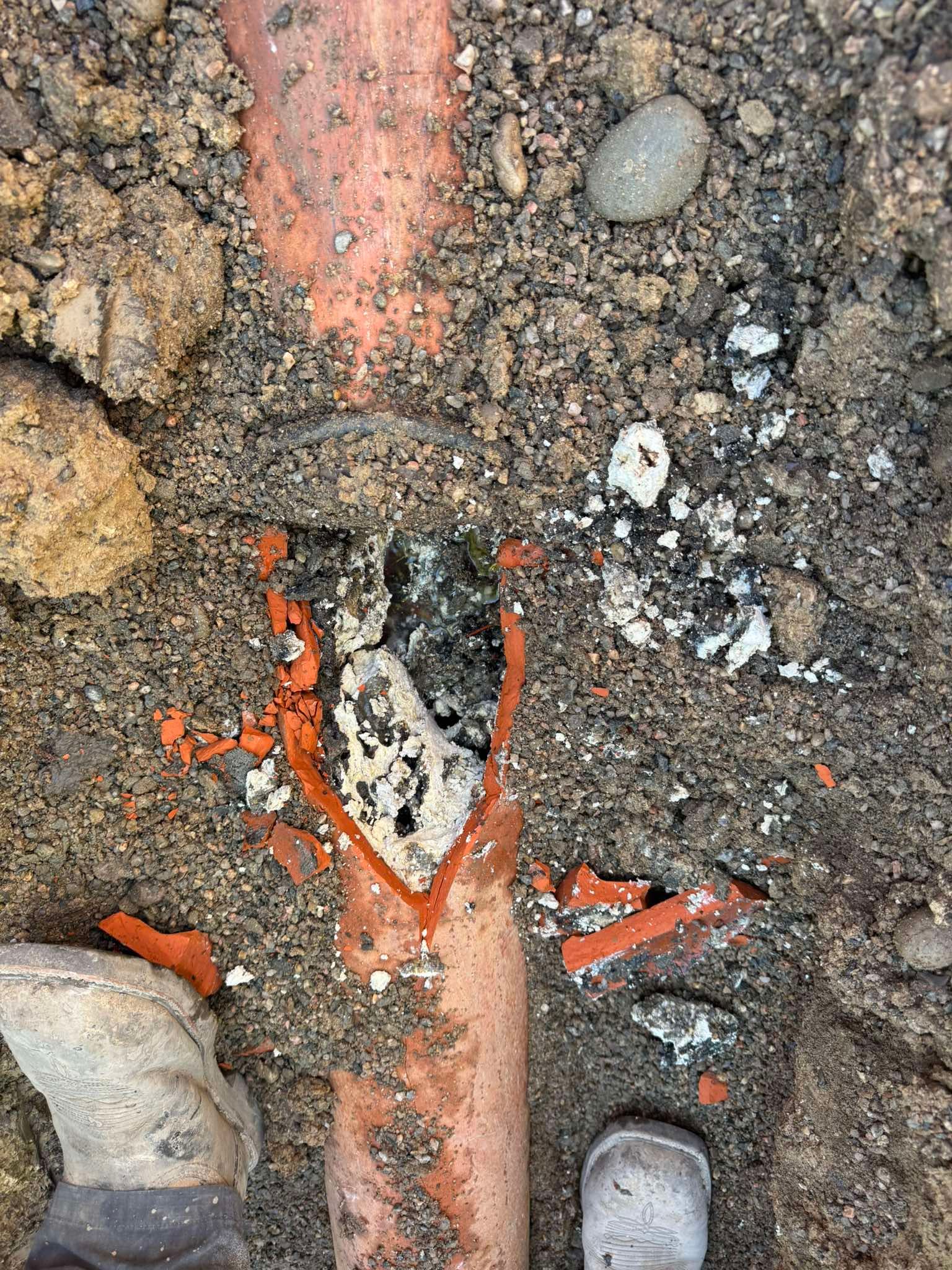 A broken orange PVC pipe partially buried in dirt, with debris and exposed interior visible at the fracture.