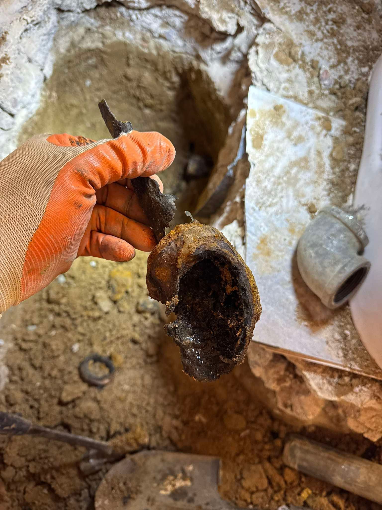 A gloved hand holds a corroded, broken pipe segment over a dirt-filled hole next to a clean pipe fitting.